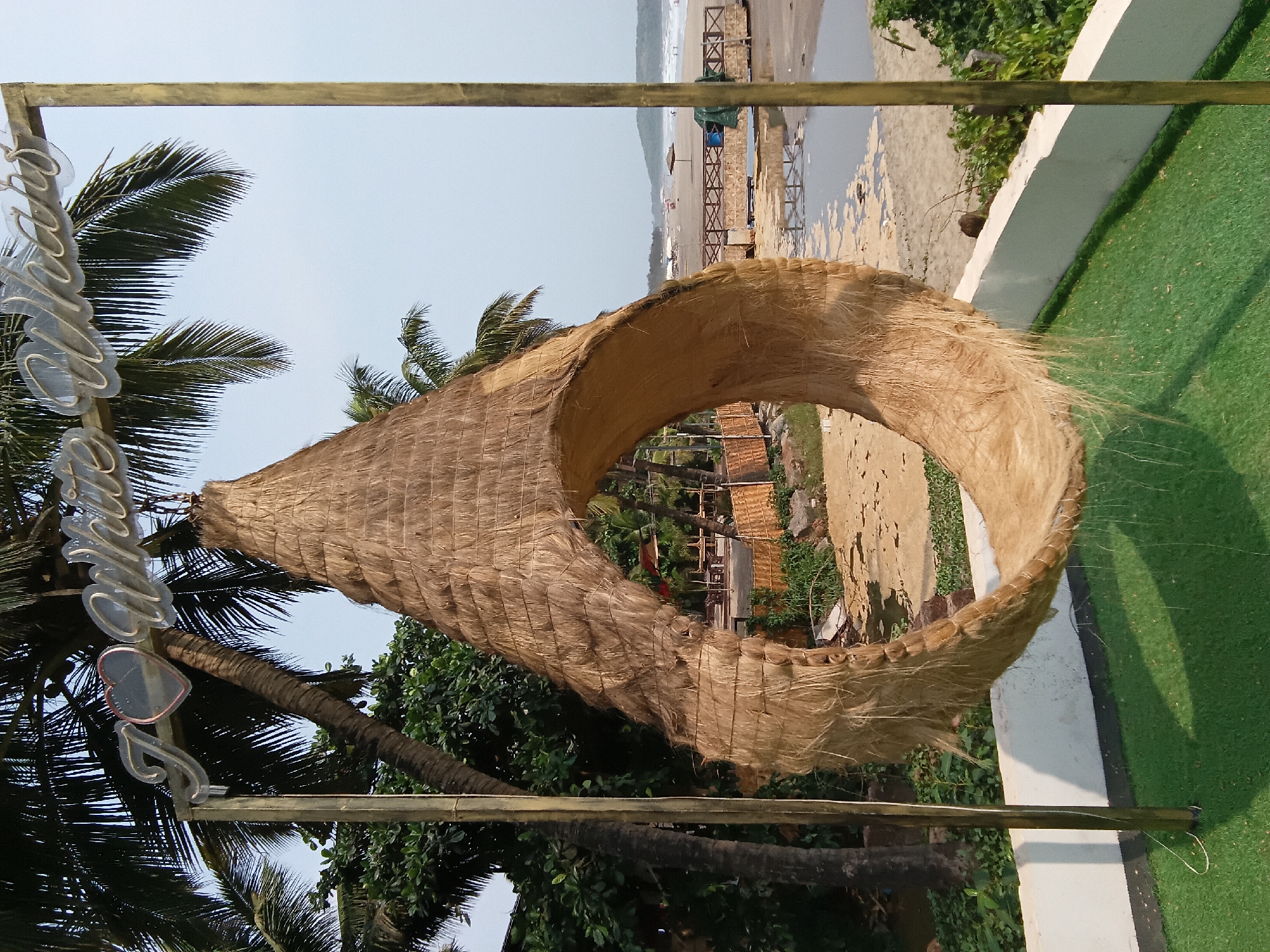The image shows a large, teardrop-shaped structure made of dried grasses or similar material, with a circular opening in the center. The structure is suspended by a simple metal frame. Through the opening, a beach scene can be observed, with sand, water, and wooden structures. Palm trees are visible in the foreground and background, lending a tropical feel. A sign with the name "Ashvem" is affixed on the left. The location is Ashvem, India.