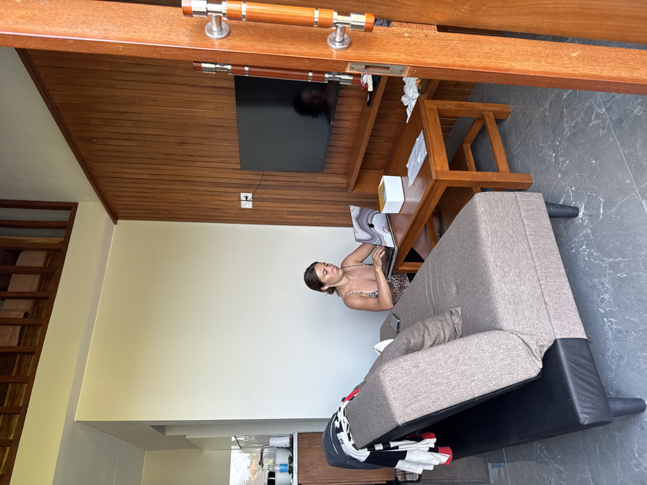 The image displays an indoor living area within a residence or accommodation located in Pecatu, Indonesia. An adult female, characterized by brown hair and wearing a leopard-print bralette or camisole, is seated on the floor to the left of a wooden coffee table. She is actively typing on an open silver laptop, which features a distinct marble-patterned skin on its lid. White wireless earbuds are visible in her ears. A smartphone rests on a gray and black two-seater sofa/futon positioned to her left, over which a striped shirt and dark garment are draped. A white square box and a piece of white paper are present on the coffee table.
The room's floor consists of dark gray tiles with a subtle marble pattern. Walls are painted light cream on the left and rear, while the right wall is covered with vertical wood paneling. A flat-screen television is mounted on the wood-paneled wall above a low wooden media console. A white power outlet with a black cable plugged in is visible on the wood wall. In the upper background, a wooden railing indicates the presence of a mezzanine level. The far left reveals a partial view of a kitchenette area, including a sink, faucet, and various containers on a counter. A partially open wooden door frame with two vertical wooden and metal handles is positioned on the far right, suggesting an entry or internal doorway. The scene is illuminated by natural light.