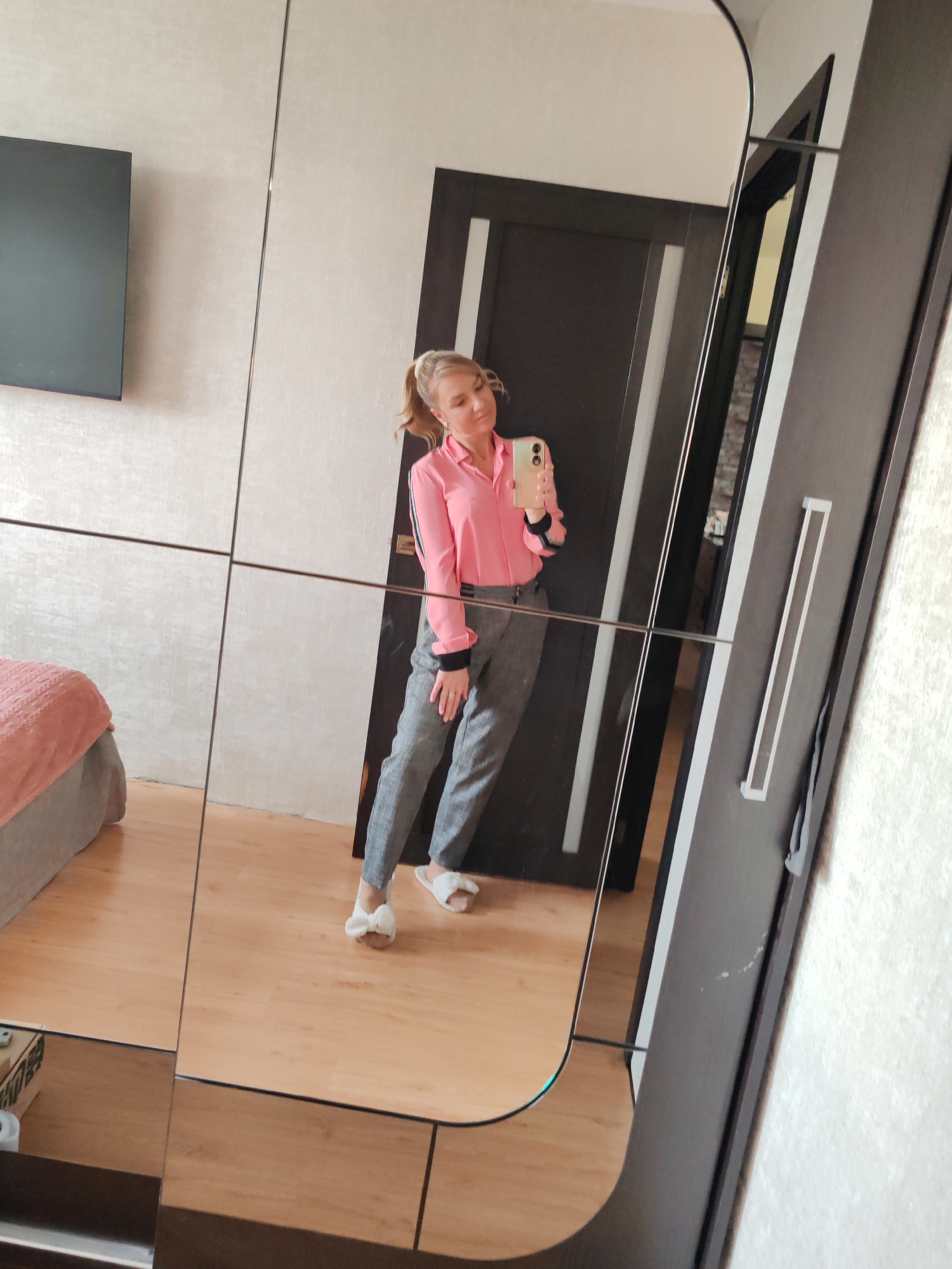 In Petropavlovsk-Kamchatsky, Russia, a young woman with blonde hair tied in a ponytail stands in a room, taking a selfie in a mirrored wardrobe. She is wearing a pink collared shirt with black stripes on the sleeves and gray plaid trousers. Her feet are adorned with white fuzzy slippers with bows. The mirrored wardrobe reflects the woman, a closed dark wooden door, a bed with a pink textured blanket, and a black rectangular object mounted on a textured wall. The room has light-colored walls and a light wood-paneled floor.