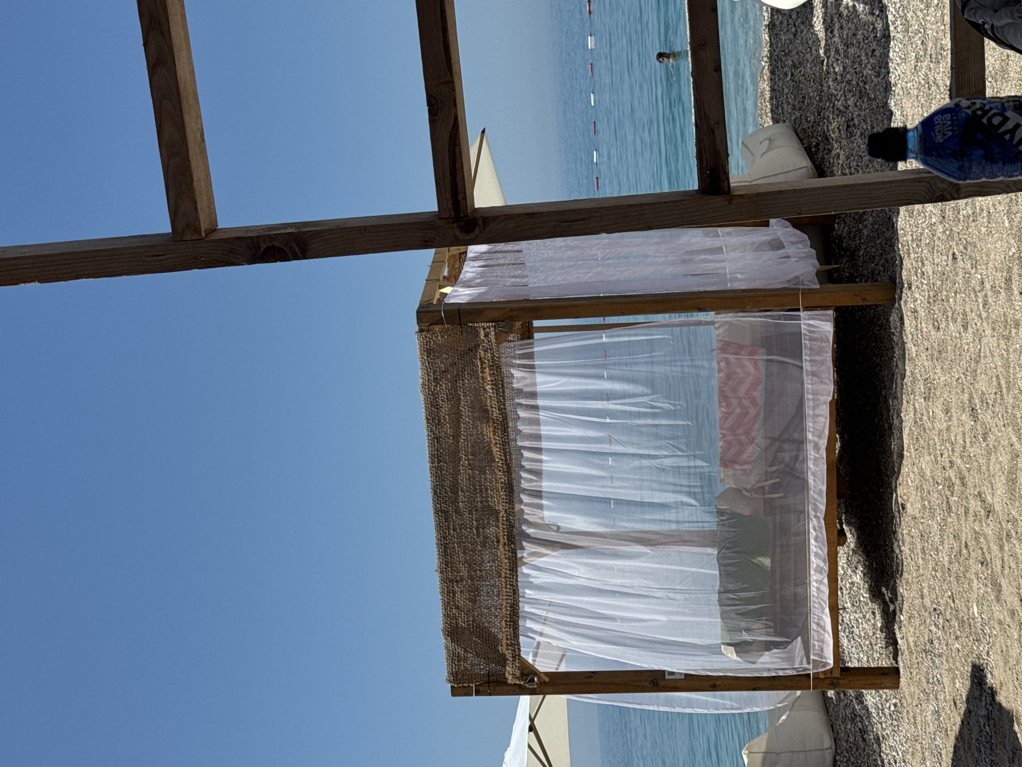The image depicts a sunlit beach scene in Vrba, Montenegro. In the immediate foreground on the right, a weathered wooden frame structure, composed of two intersecting planks, partially obscures the view. Below this structure, a blue "Aqua Viva Sport HYDRA" water bottle with a black cap rests on the pebbled beach surface.
The central focus is a wooden beach cabana or daybed, raised on four posts. Its roof is covered with a coarse, light brown woven material. White, semi-transparent sheer curtains enclose the cabana, providing partial privacy. Inside, a bed or platform is visible, furnished with a green fabric item on the left and a red and white patterned towel or blanket on the right.
To the left of the cabana, a white beach umbrella is partially visible, deployed above the horizon. A white beanbag-style cushion is situated on the pebbled beach to the left of the cabana, and another is partially visible to the right, behind the cabana. The ground consists of a mixture of sand and small pebbles/gravel.
In the background, the calm, clear blue-green sea extends to the horizon. A line of red and white cylindrical buoys is visible across the water, marking a designated area. To the right, a person's head and shoulders are visible as they swim or stand in the sea. The sky above is clear and light blue, indicating bright, sunny weather.