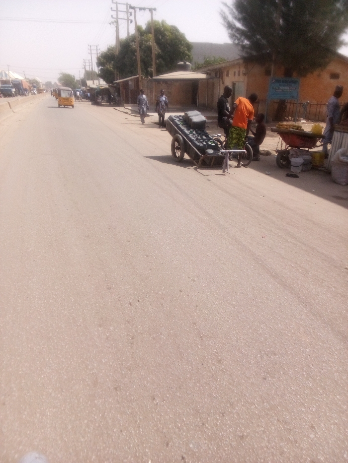 This image depicts a wide, paved road in the city of Rangaza, Nigeria, under bright daylight conditions. The road extends into the distance, appearing to be a multi-lane thoroughfare. On the left side of the road, a yellow motorized tricycle, commonly known as a Keke Napep, is moving away from the viewer.
On the right side of the road, a commercial area or market stall setup is visible. A bicycle-drawn cart, loaded with multiple dark-colored rectangular containers, is parked parallel to the road. Attached to this cart is a bicycle. Immediately behind it, a man wearing an orange shirt and a patterned green lower garment stands, facing towards the right and interacting with what appears to be a smaller individual or an object at ground level. Another man in a black shirt is standing nearby. To their right, a red wheelbarrow is filled with various items, possibly produce or goods for sale. Several white buckets and bags are situated on the ground around the wheelbarrow. Further right, a man in a light-colored top stands next to a market display.
Two men are observed walking on the right shoulder of the road, between the Keke Napep and the market stalls. Buildings with light-colored facades and various roof types, including corrugated metal, line the right side of the street. Overhanging power lines and wooden utility poles are visible above these structures and along the street. Green trees are interspersed among the buildings and utility infrastructure. The overall scene indicates active daily life and commerce along an urban street.