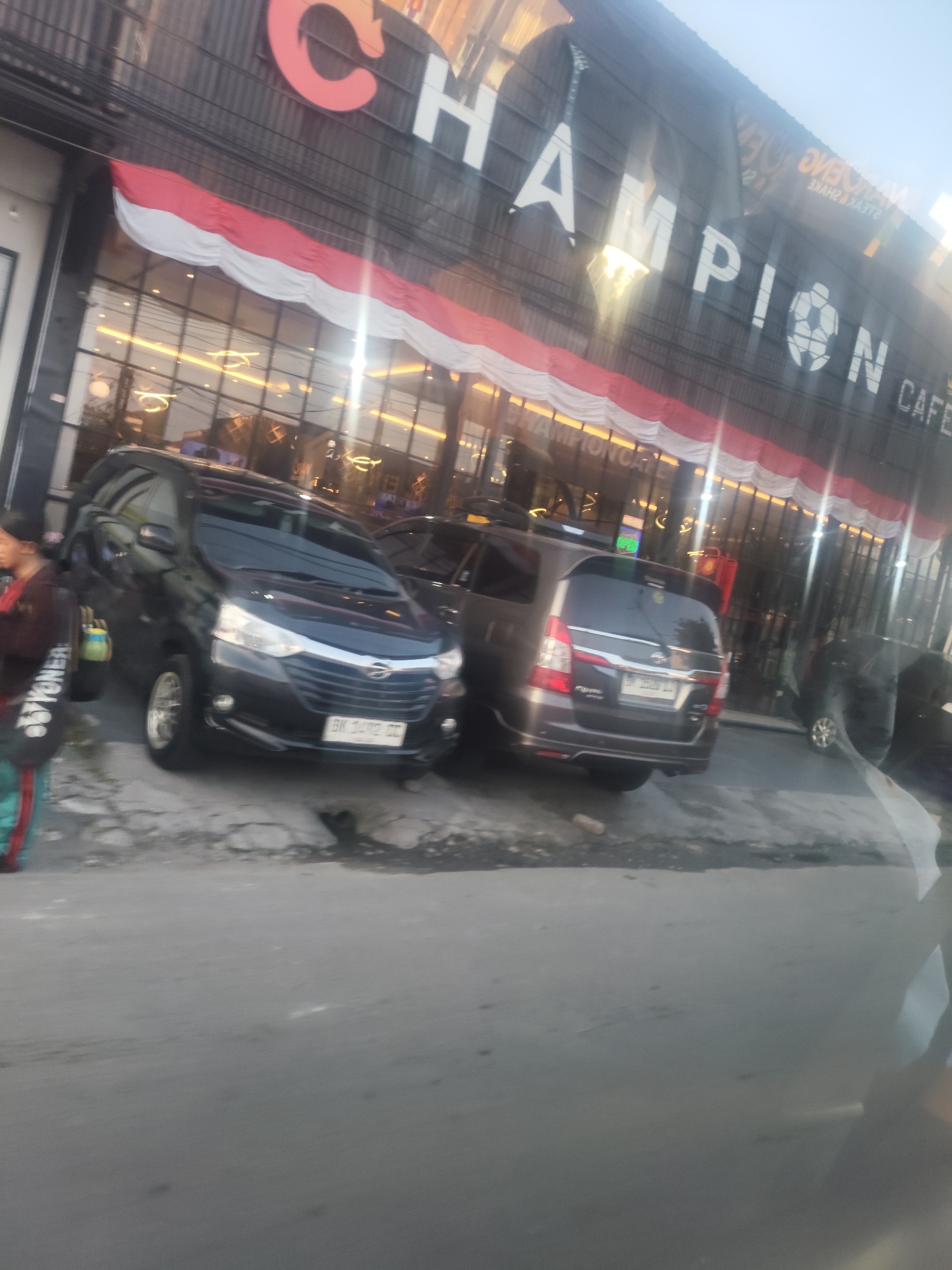 The image shows a street scene in the City of Medan, Indonesia, with a building facade featuring the word "CHAMPION CAFE" prominently displayed. The building has large windows and is decorated with a draped Indonesian flag. Two dark-colored vehicles, a sedan and an SUV, are parked on the side of the road in front of the cafe. A person is partially visible on the left side of the image, holding a bag. The overall lighting suggests it may be late afternoon or early evening.