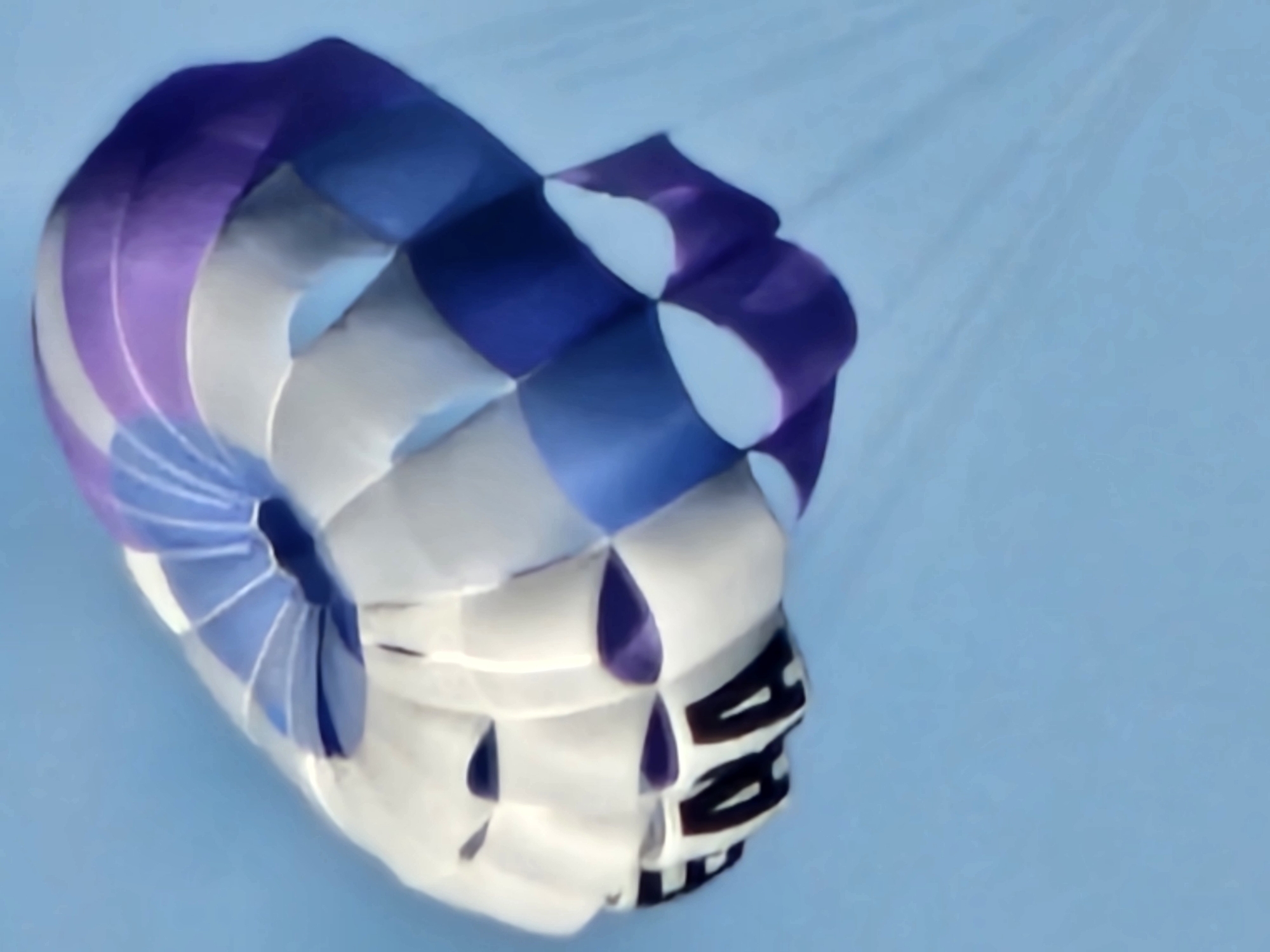 The image shows a partially deflated, multicolored parachute with visible text in the foreground, against a light blue background suggesting the sky. The parachute's color scheme consists of purple, blue, and white sections. The text on the parachute appears to be dark in color, and is located towards the lower right of the parachute's visible portion. No people are visible in the image. The environment is a clear, light blue sky, typical of a location like Kuta Selatan, Indonesia, but the lack of other features prevents confirmation of the exact location.