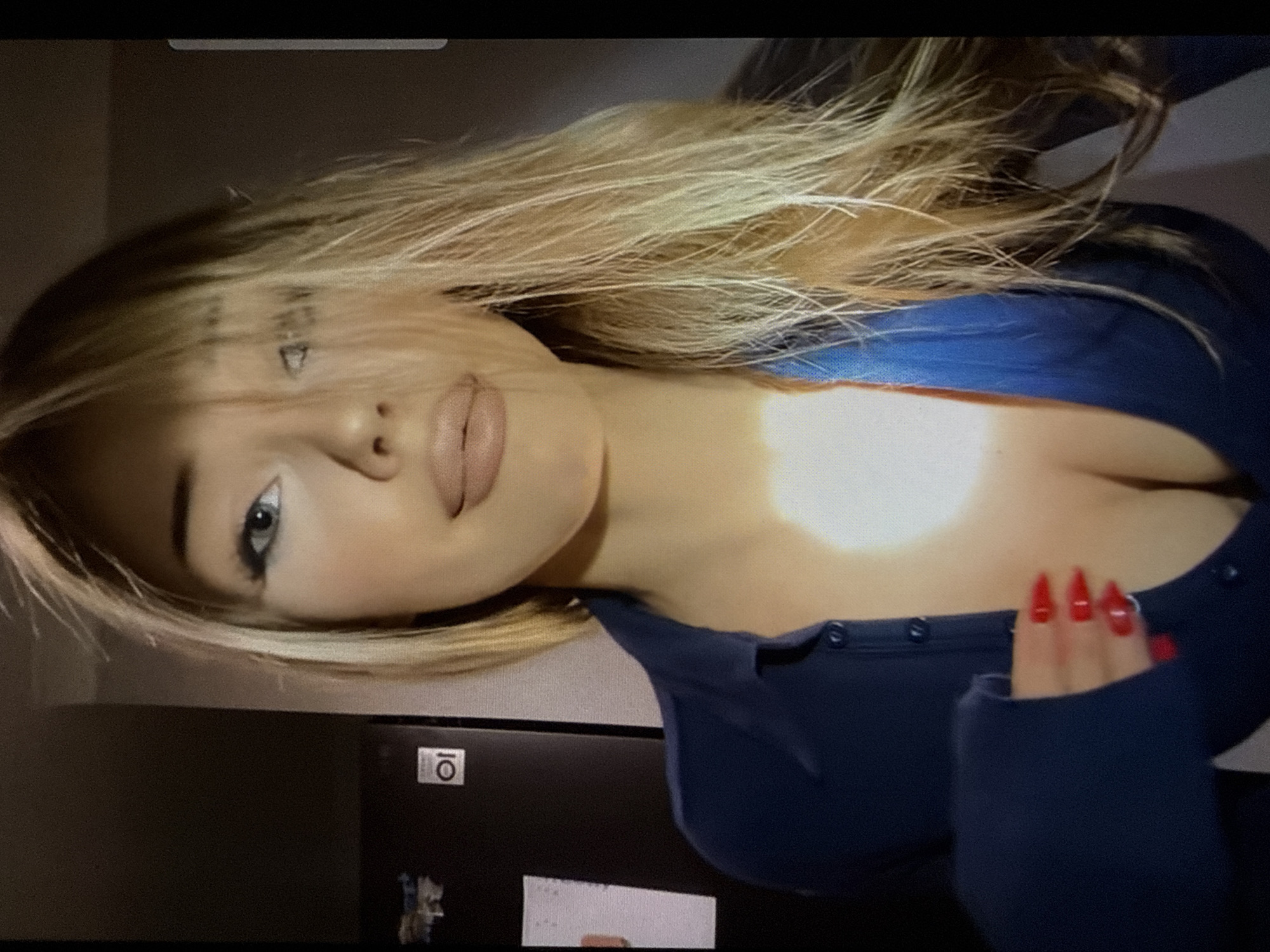 The image depicts a close-up, indoor portrait of a woman with long, light blonde hair, some strands falling across her face. She has light blue eyes with prominent winged eyeliner and full lips with light nude lipstick, exhibiting a subtle smile as she gazes directly at the camera. She wears a dark blue, long-sleeved, collared button-down top that is partially unbuttoned, revealing cleavage. Her left hand, adorned with long, pointed red-polished nails, is positioned near her chest, lightly touching the garment. The background on the left features a dark appliance, possibly a refrigerator, displaying a silver "LG" brand logo and a white circular "10" emblem. A white rectangular paper is affixed to the appliance. The right side of the background is dark and indistinct. A strong, bright light source illuminates the woman's face and upper chest, creating a significant highlight on her décolletage, contrasting with the generally dim ambient lighting of the domestic setting. The location is an indoor, domestic setting in the city of Unknown, Unknown.
