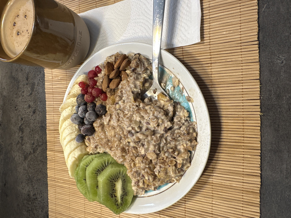 A bowl of oatmeal sits on a bamboo mat with sliced kiwi, bananas, blueberries, and red currants arranged around it. A cluster of almonds rests on top of the oatmeal. A silver spoon is partially submerged within the oatmeal. To the left, a mug of coffee complements the meal. A white paper towel is visible near the top.