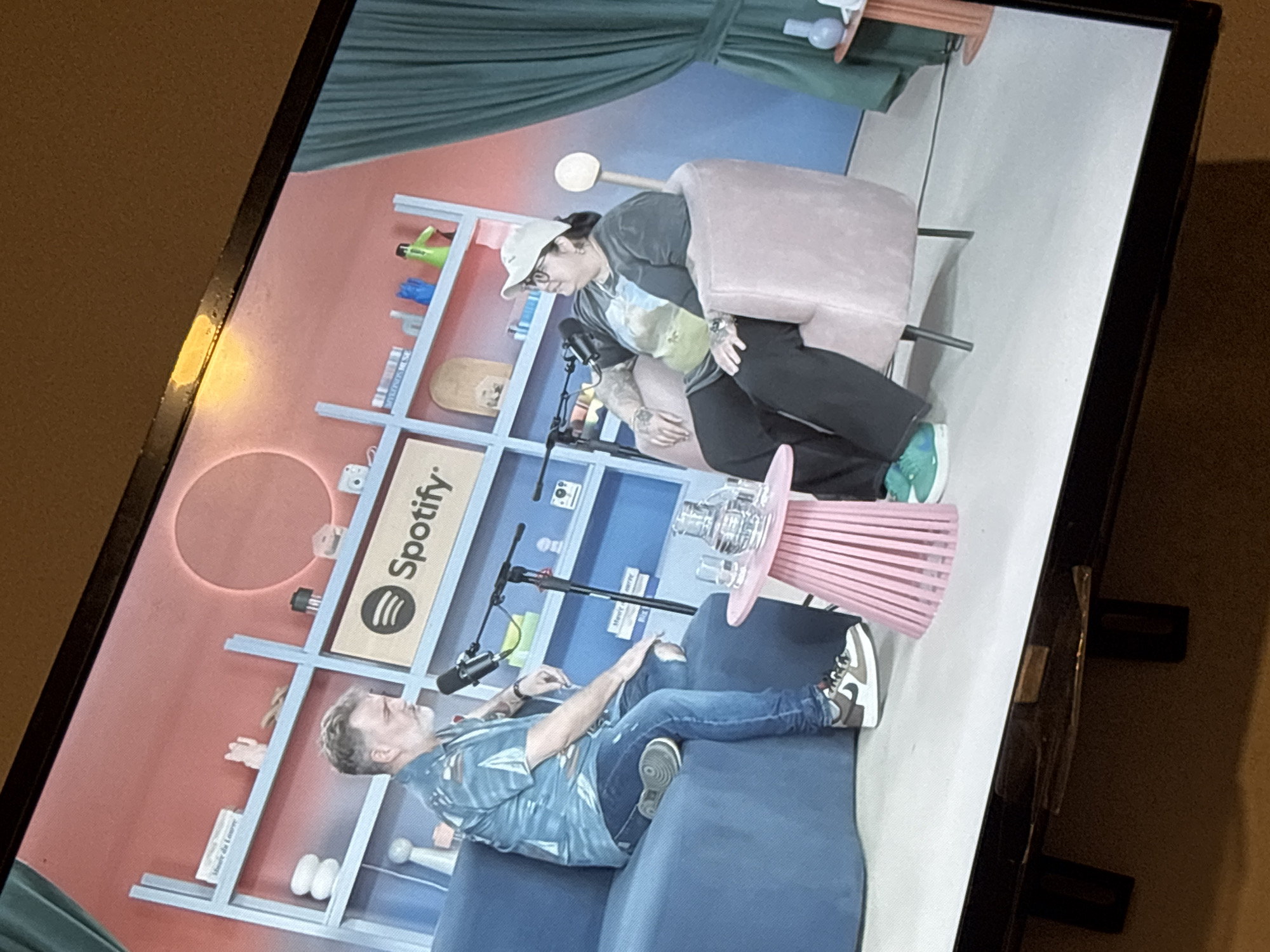 The image shows a television screen displaying a podcast recording with two people.
One person is seated on a blue sofa. They are wearing jeans and a patterned shirt.
The other person is seated in a pink armchair, wearing a baseball cap, glasses, and dark pants.
Between them is a pink side table with a clear object on top. Microphones are positioned in front of both people.
Behind them is a bookshelf and a backdrop consisting of green curtains above and a red and blue wall below. A "Spotify" logo is visible on the shelf behind the person in the armchair.
There is no information to infer the location of the scene within the city of Buenos Aires, Argentina.