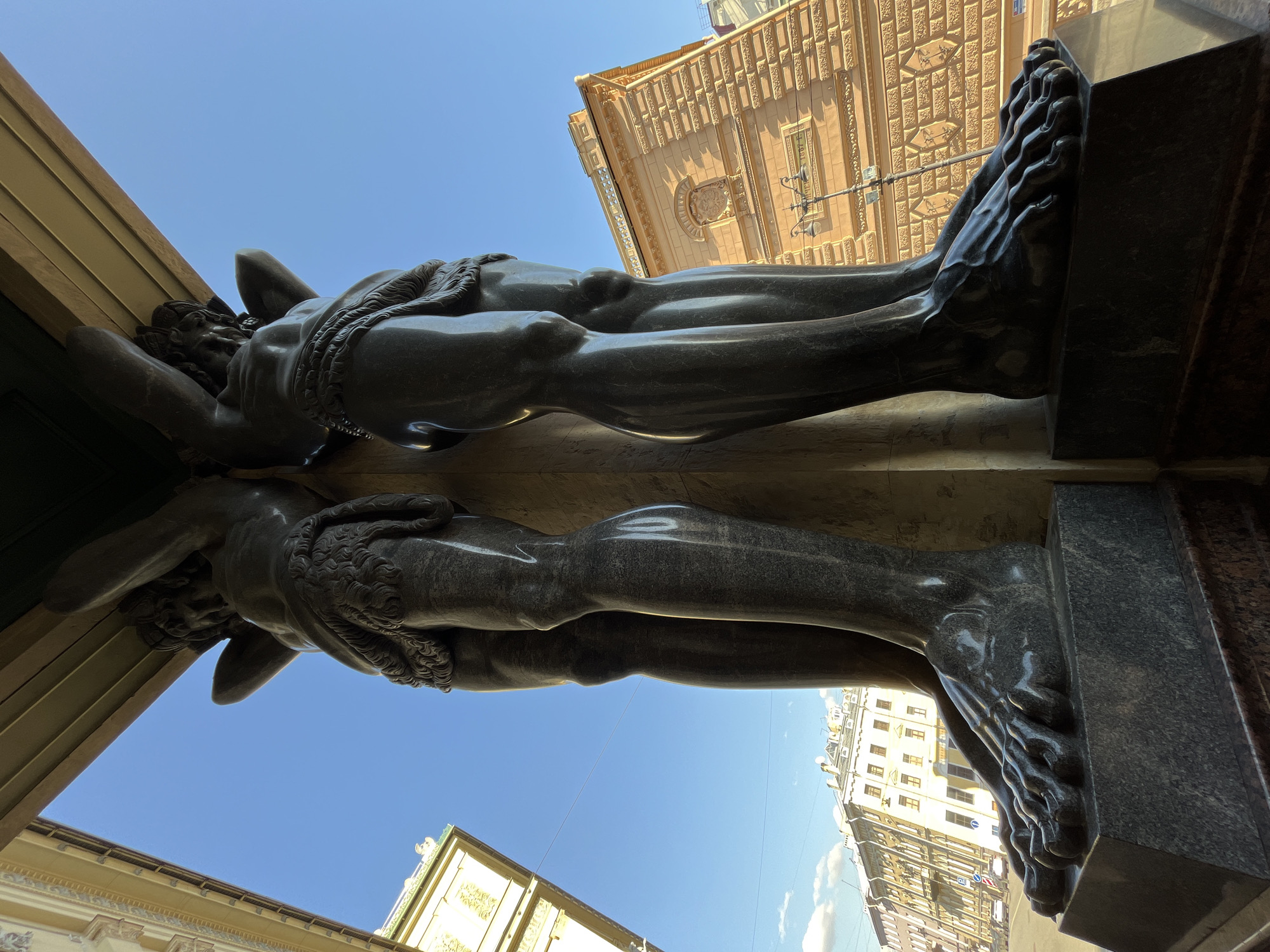 A low-angle, upward-looking view captures two dark grey granite Atlantes statues, part of the Portico of the New Hermitage building, located on Millionnaya Street in Saint Petersburg, Russia. The statues are depicted as muscular male figures, nude save for draped loincloths, with their arms raised to support a massive entablature above them, which is partially visible in the upper frame. Their polished surfaces reflect bright sunlight, emphasizing anatomical details such as strong musculature and defined feet resting on dark granite plinths.
To the left of the statues, a light yellow building with intricate architectural details is partially visible under a bright blue sky. To the right, a multi-story building with a textured, light orange facade, featuring decorative rectangular patterns and a central crest, is illuminated by direct sunlight. A black, ornate streetlamp with multiple light fixtures stands between the right Atlante and this building. In the distance, beyond the buildings, additional multi-story urban structures are visible, along with overhead power lines traversing the sky. The sky itself is clear blue with a few scattered white clouds. The scene is devoid of people or overt activity, presenting a static architectural tableau bathed in sunlight.