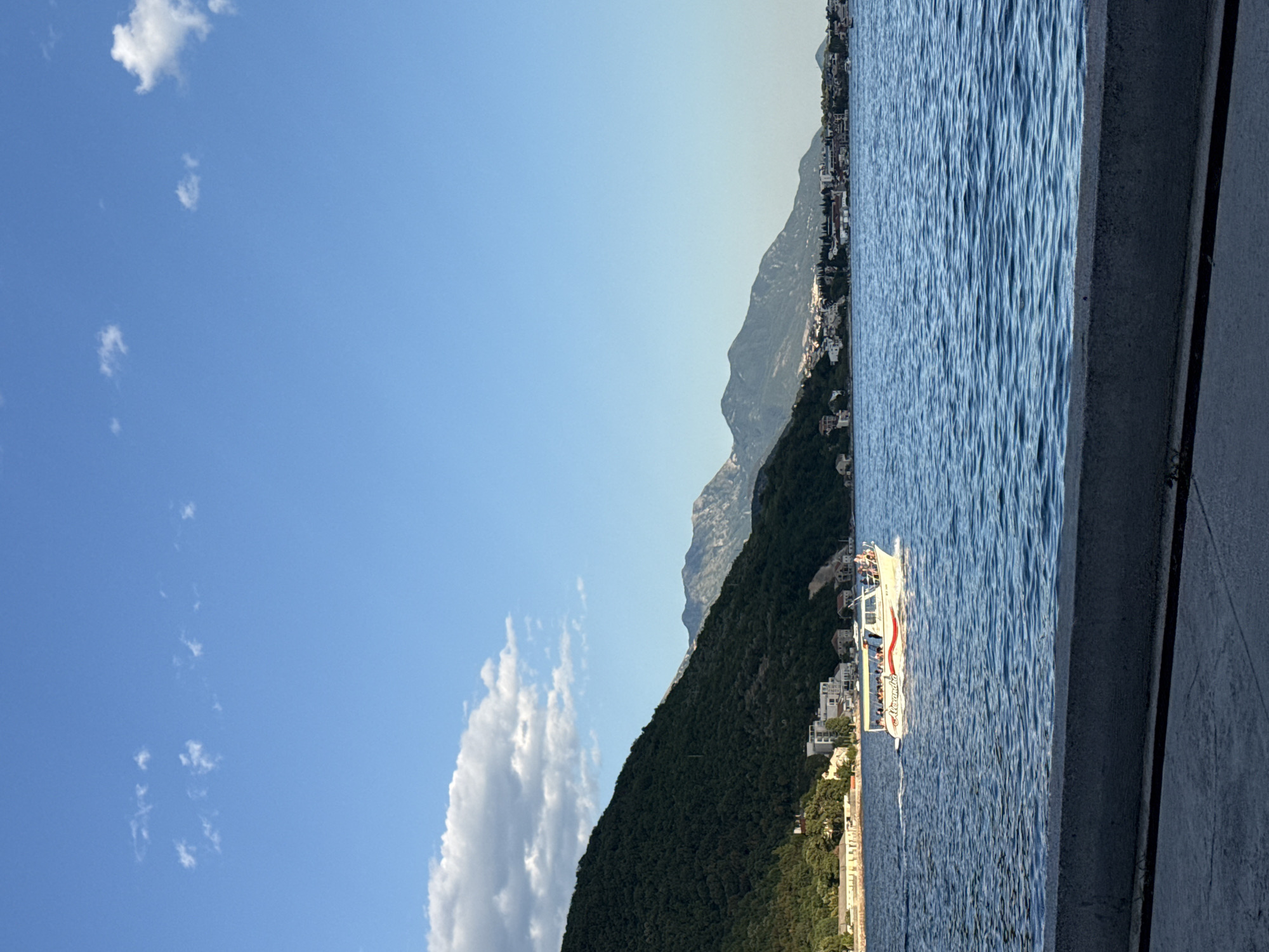 A long shot captures a picturesque scene along the coast of Jošice, Montenegro, under a clear blue sky dotted with scattered white clouds. In the foreground, a concrete barrier separates the viewer from the deep blue, slightly rippled water. A white boat with a red stripe and the name "Alexandra" emblazoned on its side is navigating the bay, carrying several passengers visible on its deck. The boat is moving from left to right.
The background features a densely forested green mountain slope rising dramatically from the water's edge, with a scattering of buildings nestled along its base and further up the incline. Beyond this, a range of larger, rugged mountains stretches across the horizon, their peaks softened by distance. A town or village is visible along the coastline, with buildings clustered together, hinting at a vibrant community. The lighting suggests it is either early morning or late afternoon, with the sun casting a gentle glow on the landscape.