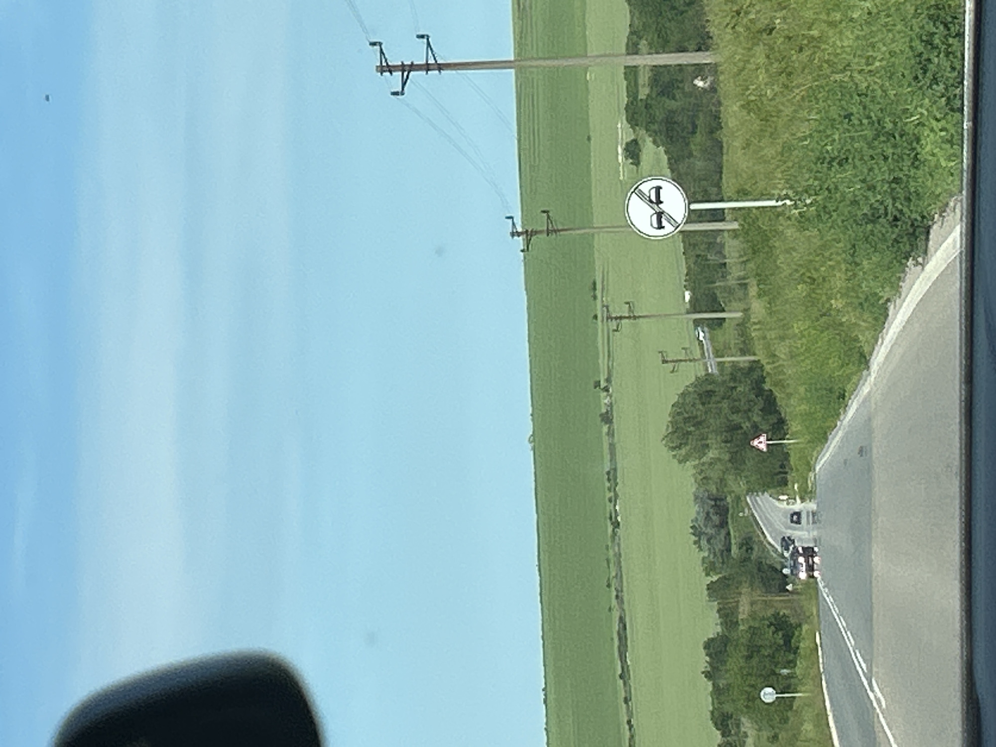 The image, taken from inside a vehicle, presents a rural road scene under a bright, clear blue sky. The perspective is rotated 90 degrees clockwise, showing the road curving gently through a countryside landscape.
On the left side of the road, extensive green agricultural fields stretch towards the horizon, interspersed with multiple wooden utility poles carrying power lines. The road itself is asphalt, marked by a solid white line separating the lanes. Ahead on the road, a line of vehicles, including what appears to be cars and possibly a truck, can be seen following the road's curve. The right side of the road is bordered by lush green vegetation.
Several European road signs are visible. A prominent circular regulatory sign features a red border with a diagonal red slash across two car silhouettes (a black car and a red car), indicating "No Overtaking" for all motor vehicles. Further down the road, a triangular warning sign with a red border and a black exclamation mark signifies "Other Dangers." Another circular sign, likely marking the end of a prohibition, is visible in the distance.
The scene depicts bright daytime conditions with clear weather. No people are explicitly visible. A dark object, possibly part of the vehicle's interior or a reflection, appears in the bottom left corner. The setting is an open countryside road, specified to be near Kumanovo, Bulgaria.