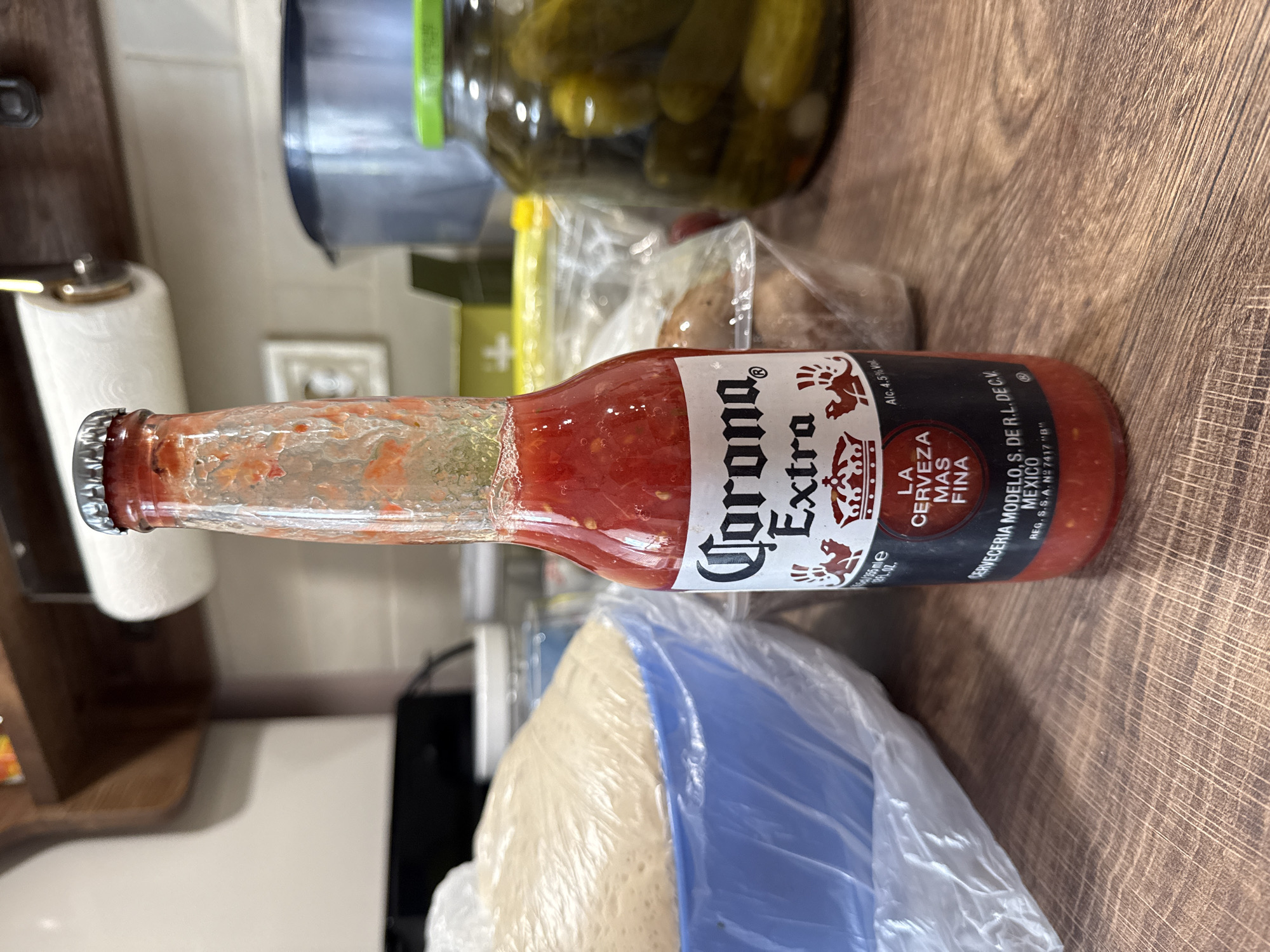 A Corona Extra bottle is lying horizontally on a wooden surface. The bottle appears to be filled with a red, pulpy substance. Other objects visible in the background include a jar of pickles, a bag of potatoes, and a bag of dough. The location is most likely a kitchen or food preparation area, not Sofia, Bulgaria.