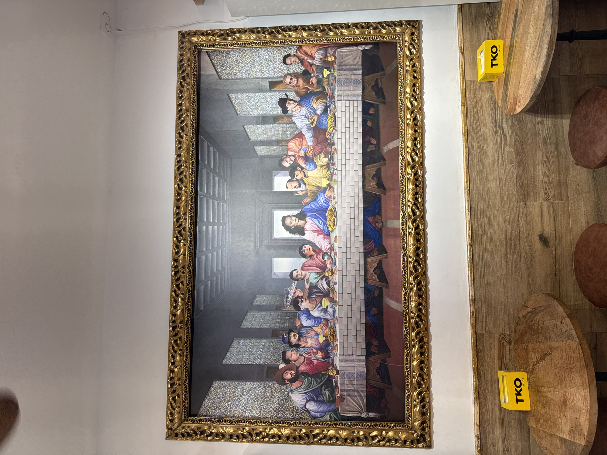 This image depicts a framed artwork, a stylized rendition of "The Last Supper," displayed on a white wall above a wooden paneled section. The painting features a group of men gathered around a table, with a central figure resembling Jesus. The men are dressed in various modern attire and accessories, including hats and sunglasses, and some are holding what appear to be tacos. The room is lit, and the artwork is framed in an ornate gold frame. In the foreground, two round wooden tables are visible, each with a yellow "TKO TACOS" box on it. The flooring is wooden planks. This scene is located in Valencia, Spain, at a restaurant called TKO Tacos.