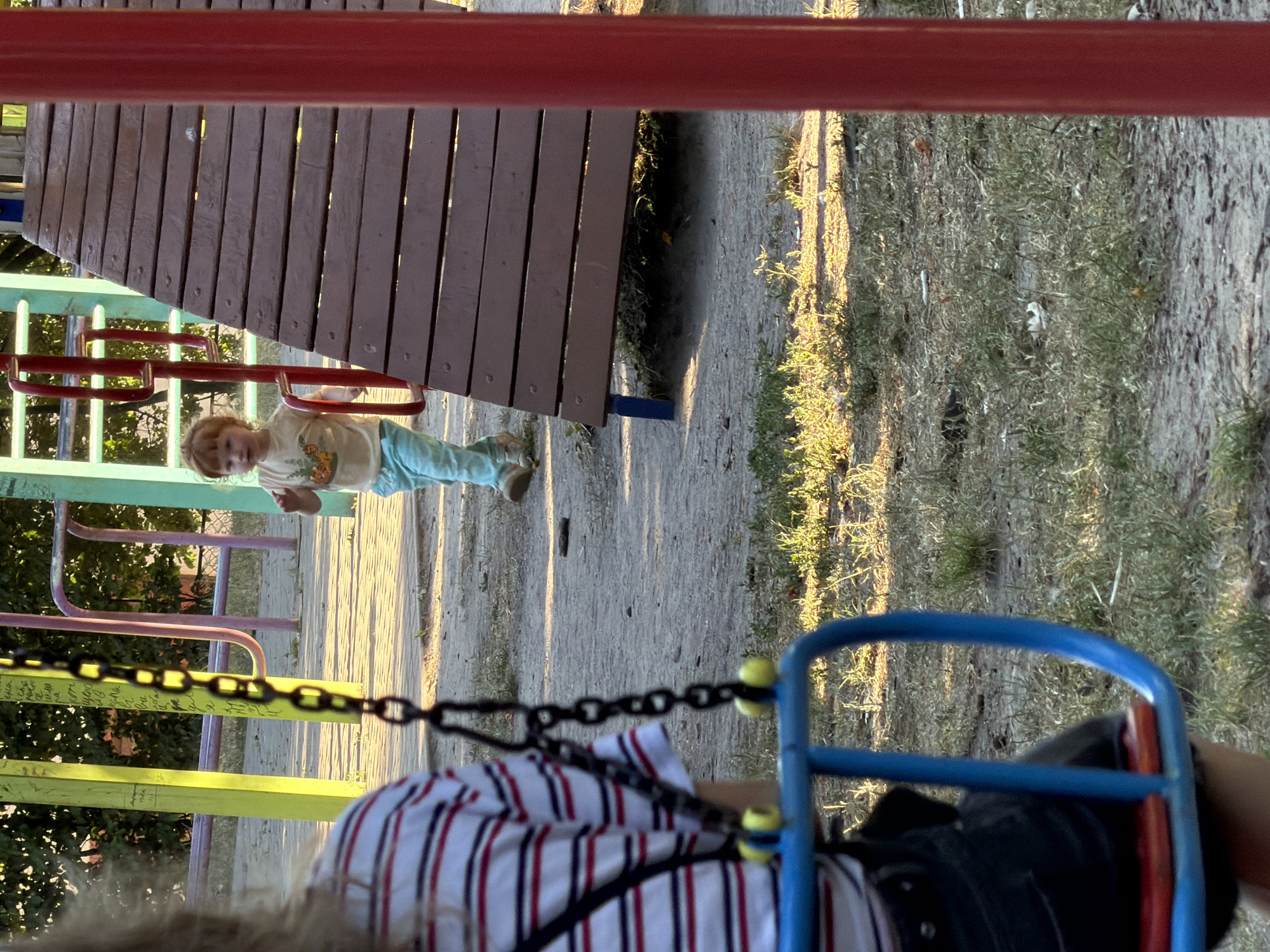 The image shows a playground scene in Kamianka-Buzka, Ukraine. The perspective is from someone on a swing, looking towards the playground structure. The time of day appears to be late afternoon, with long shadows cast across the sandy ground and dry grass.
In the mid-ground, a young child with reddish-blonde hair is standing on a wooden platform, looking towards the camera. The child is wearing a light-colored t-shirt with an animal graphic and light blue pants. The playground structure features various colored metal bars and a wooden deck, suggesting it is a well-used area. The swing set chains are prominently in the foreground, and the back of another person is visible, also appearing to be on a swing.
The overall mood is relaxed and informal, typical of a playground on a sunny day. The dry vegetation suggests a warm season. There is no visible text in the image.