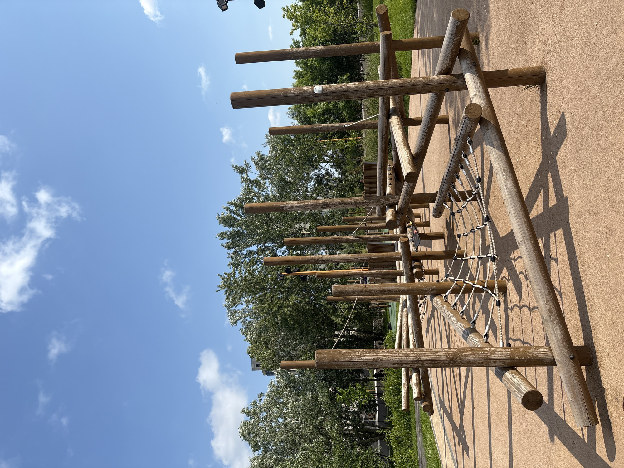 The image shows an outdoor children's playground, likely in Saint Petersburg, Russia, due to the architectural style of the distant buildings and the type of vegetation, which is common in the region. The playground features a complex wooden climbing structure with numerous logs and ropes. A net made of thick white ropes is suspended between some of the wooden elements, serving as a climbing net or a bridge. The ground surface is a reddish-brown, likely a soft material for safety, such as rubber mulch or compacted sand. In the background, several trees with green foliage line the perimeter of the playground. Behind the trees, a tall, modern building with a light-colored facade is visible, suggesting an urban or suburban setting. The sky is a clear, bright blue with a few scattered white clouds. The lighting suggests it is daytime, with the sun casting distinct shadows from the wooden structures onto the ground. There are no people visible in the immediate vicinity of the playground structure in the foreground, though a figure can be seen in the mid-ground, walking away from the camera in the park area.