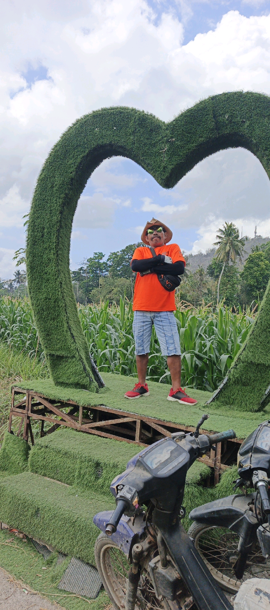 An outdoor medium shot captures a man standing with his arms crossed in front of a large green heart-shaped topiary. The man is wearing a straw hat, sunglasses, an orange t-shirt, and blue shorts. He has a mustache. He is positioned on a wooden platform covered in artificial grass, which is part of the heart structure. Behind the heart, a field of corn is visible, and in the distance, there are trees, hills, and a cloudy sky. In the foreground, two motorcycles are parked, partially obscuring the base of the platform. The scene is likely a tourist attraction.