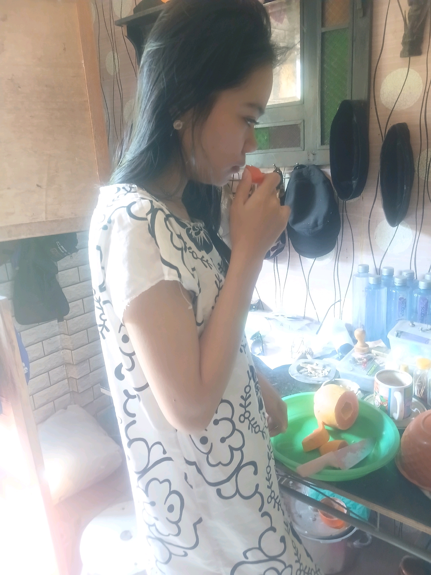 A young woman with long, dark hair is standing in what appears to be a kitchen or food preparation area. She is wearing a white dress with a black floral pattern. Her right hand is raised, holding a small piece of orange fruit, possibly a papaya, to her lips. In front of her, on a green plastic plate, are pieces of peeled papaya and a knife, suggesting she is preparing to eat or has been cutting the fruit. Behind her, there are shelves with various items, including several plastic bottles of water, a mug, and black hats hanging on the wall. The background shows a patterned wallpaper and a window. The overall impression is a candid moment of someone casually preparing or eating fruit in a domestic setting. There are no specific visual cues to indicate the location in Banyuwangi, Indonesia, beyond the initial context provided.