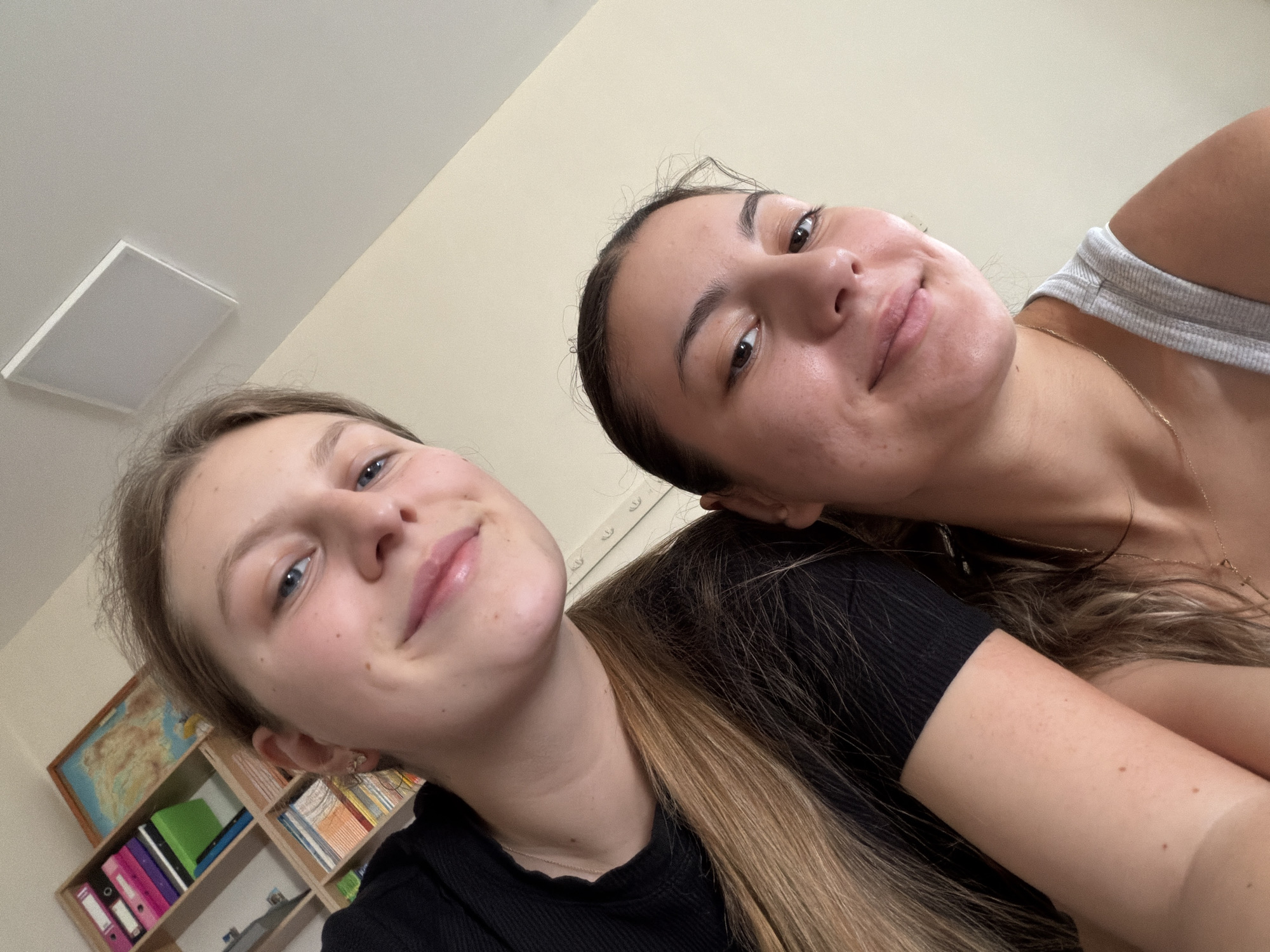 The image shows two young women in a brightly lit room. The woman on the left, with blonde hair, is wearing a black top and is positioned slightly behind and to the left of the other woman. She has blue eyes and light skin with some freckles. The woman on the right, with dark hair, is wearing a grey tank top. She has brown eyes and her skin appears to have some blemishes. Behind them, a wooden shelf unit is visible, filled with books and folders of various colors, including pink and blue. A map is also displayed on the shelf. The ceiling above them is white, and a square light fixture is mounted in the upper right corner. The overall environment appears to be domestic, possibly a bedroom or study area. There is no identifiable information suggesting the location is in the city of Burgas, Bulgaria.