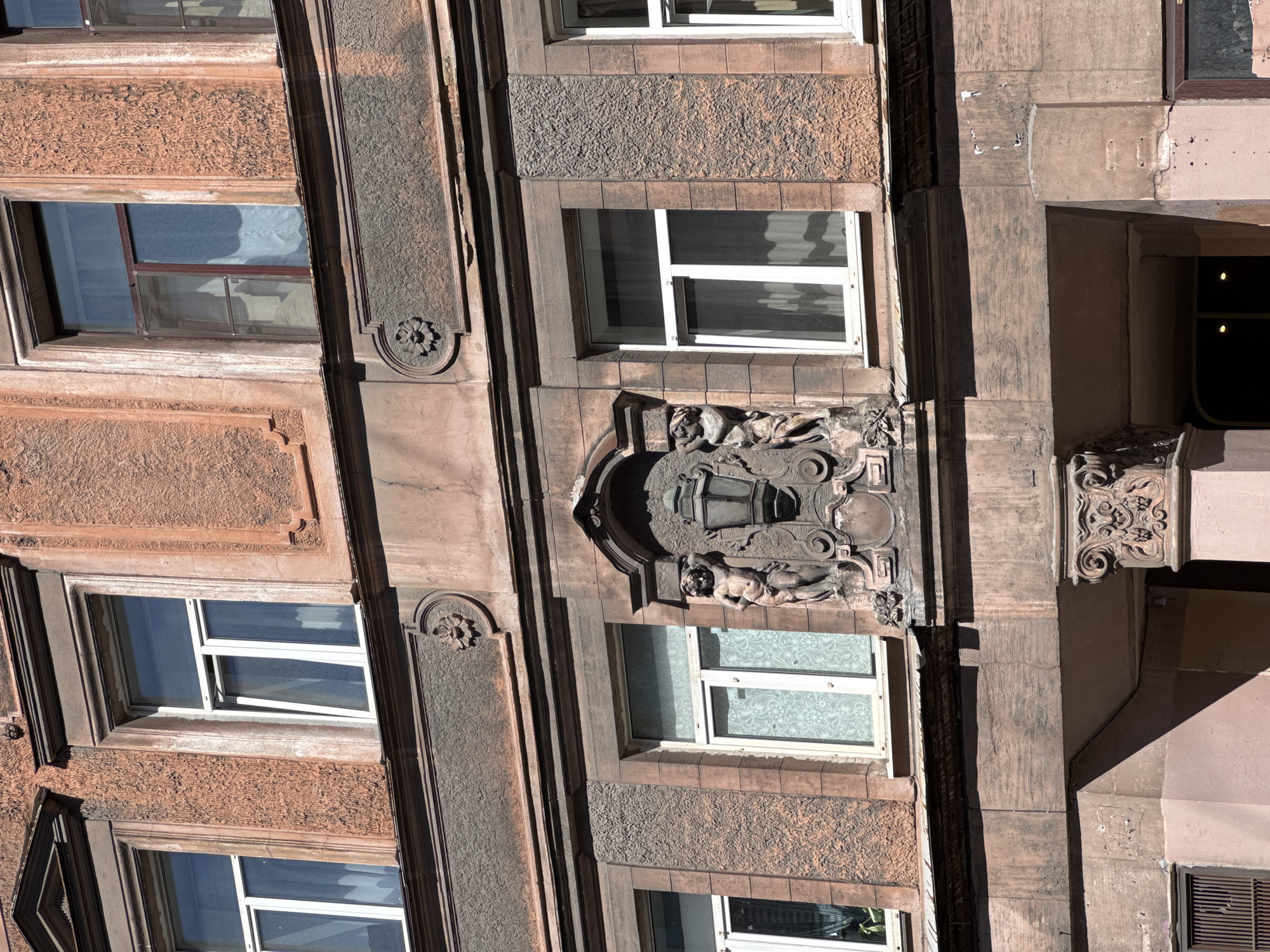 This image captures a detailed close-up of the facade of a historic multi-story building located at Malaya Morskaya Ulitsa 12, Saint Petersburg, Russia. The building exhibits architectural elements characteristic of early 20th-century eclecticism or Art Nouveau.
The facade is primarily composed of reddish-brown and muted pink stucco with varying textures, including roughcast sections and smoother, darker horizontal bands and window surrounds. The central focal point is an elaborate sculptural composition positioned above an arched entranceway, the upper part of which is visible at the bottom. This central element features a large, ornate lantern or lamp, depicted in a stylized, somewhat classical manner. It is flanked by two nude male figures, resembling atlantes or herms, who appear to support the structure of the lantern. This entire sculptural group is set within a decorative, arched niche.
Above this central sculpture, a darker frieze-like band runs horizontally, adorned with circular relief medallions depicting floral rosettes at regular intervals.
Multiple rectangular windows are visible across two floors. On the upper floor, windows have a mix of dark and modern white frames. The window on the far right of the upper floor has dark frames, while the one to its left, partially visible, has a white frame and is slightly ajar. On the middle floor, the windows to the left and right of the central sculpture both have modern white frames. Various curtains or blinds, some with floral patterns, are visible through the glass.
The building's surface displays signs of age and wear, including minor cracks, patches of discoloration, and a weathered appearance on the stucco and decorative elements. Below the central sculpture, on the right side of the entrance, a highly ornate column capital is visible, featuring intricate volutes and what appears to be a carved grotesque face or mask. Strong sunlight from the upper left casts prominent shadows across the facade, accentuating its three-dimensional details and textures, indicating a bright, clear day. No people are present in the scene.