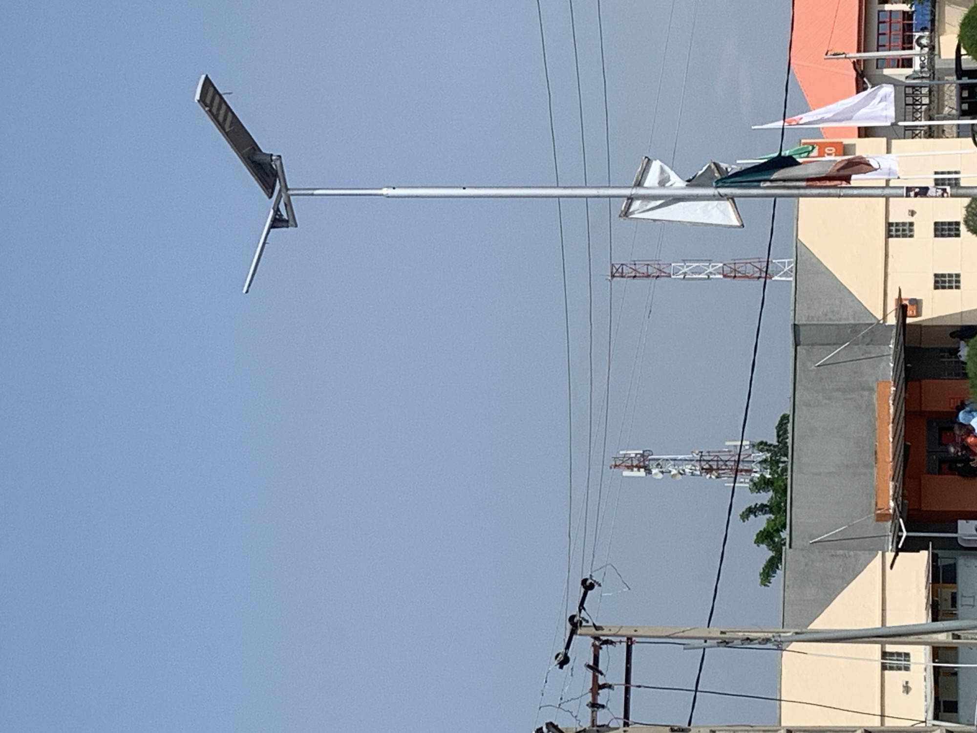 A bright, clear sky is visible in the background of an outdoor scene. In the foreground, a tall, silver lamppost with a solar panel on top stands prominently. To the left of the lamppost, there is a utility pole with wires. Further in the background, two cell towers can be seen, one red and white and the other white and red. Several buildings with orange and beige facades are also present, along with some trees. A few people are visible near the entrance of one of the buildings. There are also flags displayed on a pole near the lamppost.