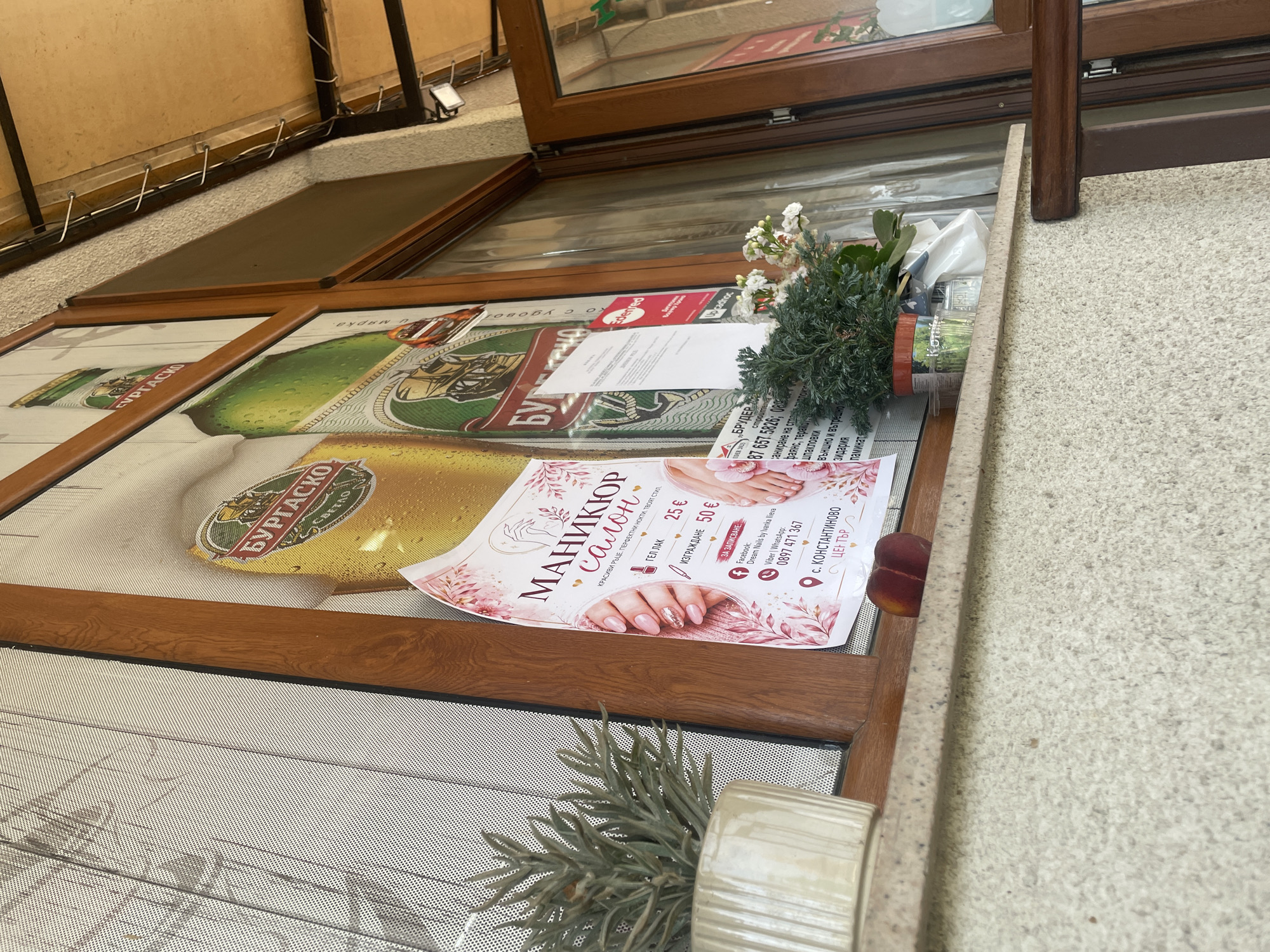 The image displays an eye-level, slightly upward-angled view of a building's exterior, focusing on a window display and adjacent architectural elements in Konstantinovo, Bulgaria.
The central feature is a window with dark brown wooden frames. The left-most pane is covered by a fine mesh screen, partially obscuring the view through it. A light-colored, ribbed ceramic pot containing a spiky green plant is positioned on the windowsill on the left side.
Several posters are affixed to the windowpanes. A large advertisement for "БУРГАСКО" (Burgasko) beer is visible, depicting a glass of beer with foam and a green beer bottle. Overlaid or adjacent to this is a pink and white poster for a "МАНИКЮР салон" (Manicure Salon). This poster illustrates manicured hands with pink and glitter nails, and a pair of feet with pedicured toenails. The text details services: "ГЕЛ ЛАК" (Gel Lacquer) for 25 € and "ИЗГРАЖДАНЕ" (Build/Extension) for 50 €. Contact information includes a Facebook page "Dream Nails by Nenka Ilieva" and a Viber/WhatsApp number: 0897 471 367. The salon's location is specified as "с. КОНСТАНТИНОВО ЦЕНТЪР" (village Konstantinovo, Center). To the right of the manicure poster, several white papers and a red card with the "Edenred" logo are partially visible, also attached to the window.
On the windowsill, between the two potted plants, two reddish-purple peaches or nectarines are resting. To the right of these fruits, another potted plant with dense green foliage and small white flowers is present in a dark pot. A crumpled white item is located behind this plant.
The wall beneath the windowsill features a light-colored, granular, textured finish, consistent with stucco or pebbledash. Above the window and extending to the right, a beige-colored ceiling or canopy structure is visible, with multiple metal hooks and electrical wiring running along its edge. A small, white, rectangular outdoor light fixture is mounted on the wall below this canopy, to the right of the window frame. An open wooden-framed door is partially visible on the far right, revealing a darker interior space.