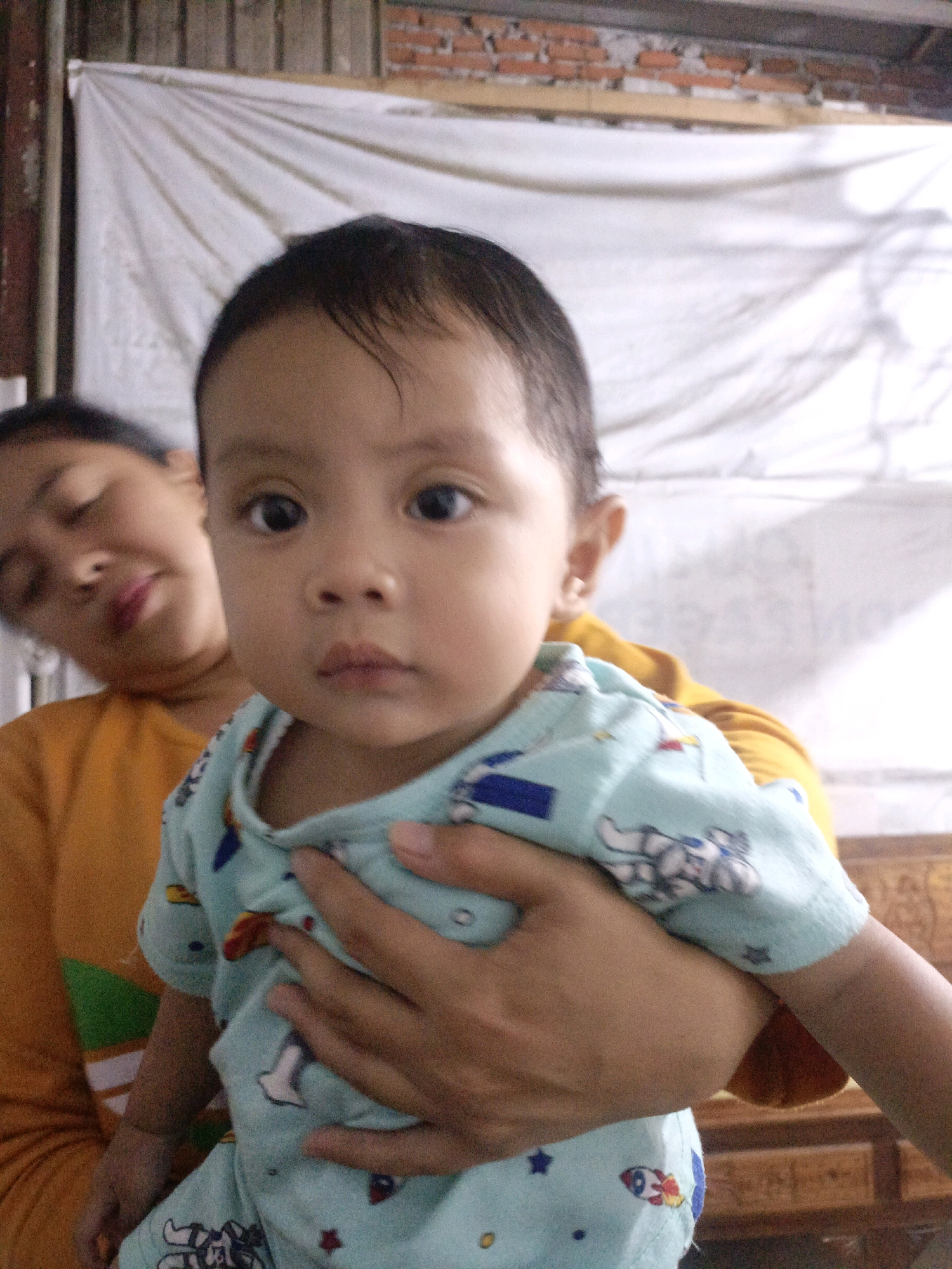 A close-up, waist-up shot shows a baby being held by a woman. The baby, in the foreground, is looking directly at the camera with a neutral expression. The baby is wearing a light blue onesie with a cartoon pattern of astronauts and stars. The woman, in the background, is out of focus, with her head tilted and eyes closed, appearing to be asleep or resting. She is wearing an orange shirt. The background consists of a white sheet hanging behind them, with a brick wall visible at the top and a wooden surface at the bottom. The overall lighting is soft. This image appears to have been taken indoors.