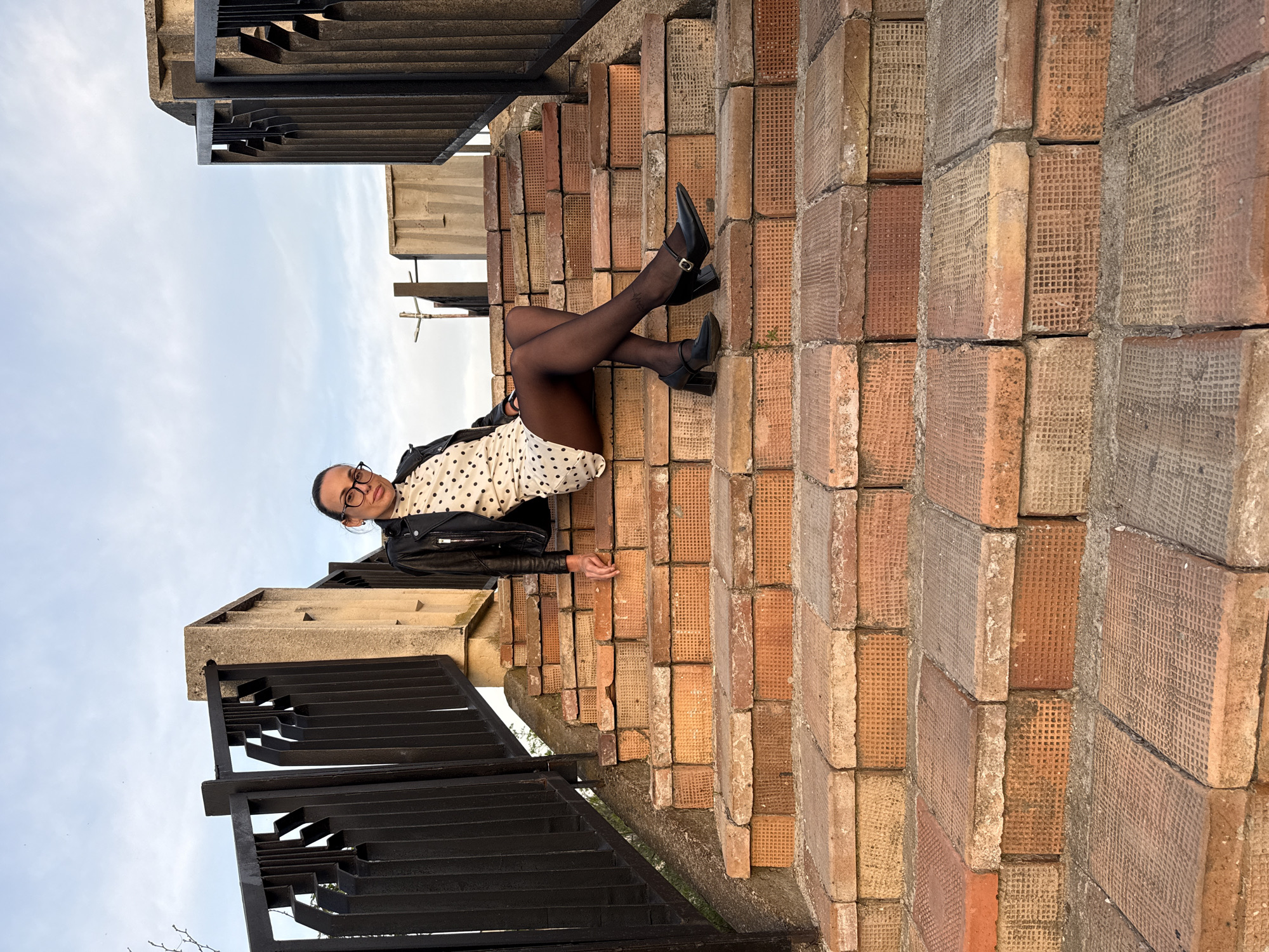 An upward-angled shot captures a person, likely a woman, posing on a terraced brick structure against a clear sky in Pleven, Bulgaria. The person is reclining with their back to the textured brick wall/stairs, looking towards the camera. They wear a black leather jacket over a white long-sleeved top with black polka dots, black sheer tights, and black high-heeled shoes with a strap across the instep. A black fanny pack or belt bag is visible around their waist. They are wearing glasses.
The background consists of light blue sky with some soft clouds, indicating daytime, possibly late afternoon due to the gentle lighting. There are no harsh shadows, suggesting overcast conditions or diffused light. Adjacent to the person and extending into the foreground are dark, geometric metal railings or a structural element, possibly part of an outdoor staircase or architectural feature.
The brickwork is a prominent detail, featuring individual bricks with a distinct textured, grid-like pattern on their visible faces. The bricks vary in shades of orange, red, and beige, with visible mortar lines. The overall setting appears to be an outdoor public space or a building's exterior, characterized by its unique and aged brick construction and modern metal accents. There is no visible text in the image.