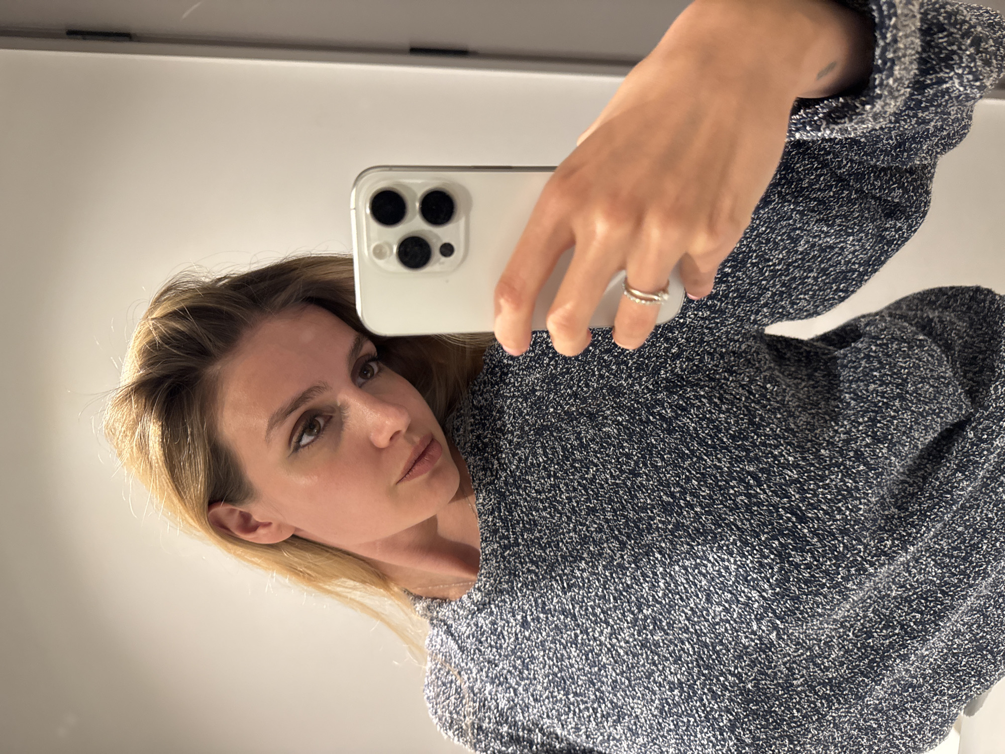 This media file captures an indoor mirror selfie of a woman. The image is rotated 90 degrees clockwise.
**Subjects:** The primary subject is a woman with blonde or light brown hair and light brown eyes. Her right hand, holding a smartphone, is also prominent.
**Objects:** A white or light-colored smartphone with a triple-lens camera array is held by the woman. She wears a silver-toned, multi-band ring on her right ring finger. Her attire consists of a dark blue or grey marled knit sweater with white flecks.
**Scene, Setting, and Activity:** The scene is an indoor setting, likely a room with a plain, light-colored wall or surface reflected in the mirror. The activity taking place is the woman taking a mirror selfie of herself with her smartphone.
**Notable details:** The woman has a neutral to slightly contemplative expression, looking directly towards the phone's lens. A small, faded tattoo is visible on her left forearm, just above the cuff of her sweater. The lighting appears soft and even, typical of indoor illumination. There are no distinct location cues beyond the implied interior space.
**Visible text:** There is no visible text in the image.