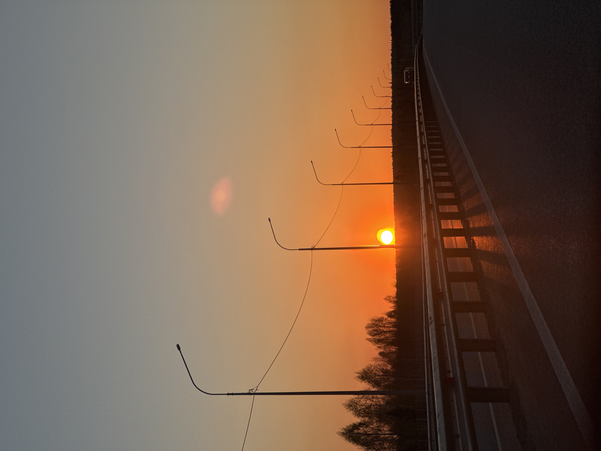 The image depicts a sunset over a road in Ivanovka, Russia. A guardrail separates the road from a line of trees. Streetlights line the side of the road, with the sun appearing to be positioned behind one of them. The sky transitions from a dark blue-gray at the top to an orange-yellow near the horizon. A vehicle is visible further down the road.
