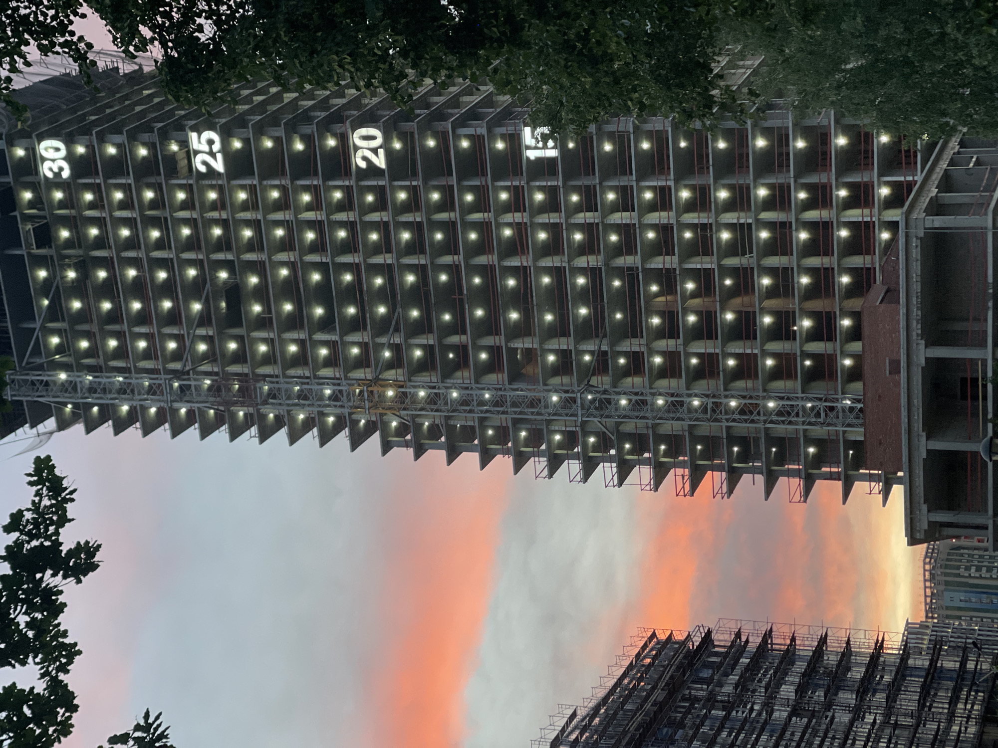 A tall, unfinished concrete structure, likely a building under construction, dominates the scene, appearing to be in the city of Izhevsk, Russia. The building has open floor spaces illuminated with lights, each one placed inside the structure's framework. Numbered signs, "30," "25," "20," and "15," are visible on the upper section of the building, possibly indicating floor levels. The structure is partially obscured by trees, and another building under construction appears in the lower part of the image. The sky exhibits pink and orange hues, suggesting sunset or sunrise.