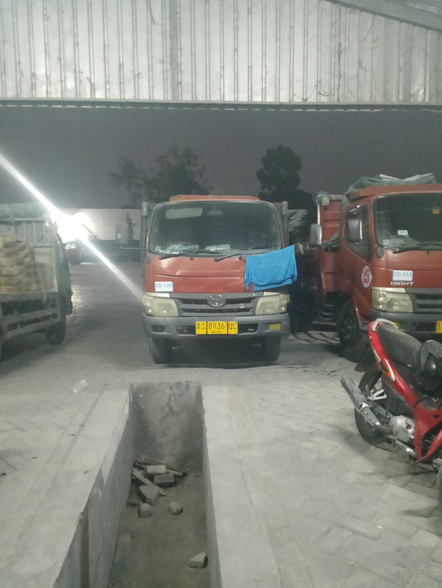 The image depicts an outdoor scene at night, likely in a parking lot or workshop area, in the vicinity of Jambean, Indonesia. In the foreground, a red motorcycle is parked on the right. The main subjects of the image are two red trucks. The truck on the left has a blue cloth draped over its front. The truck on the right has a logo with a circular emblem. The trucks are parked on a concrete surface. To the left of the trucks, another truck is partially visible, loaded with what appears to be sacks of material. In the middle ground, there is a recessed concrete pit containing debris, possibly bricks and stones. The background reveals a corrugated metal structure, possibly a building or a wall, with some trees visible in the distance. The lighting suggests artificial illumination from the overhead structure.