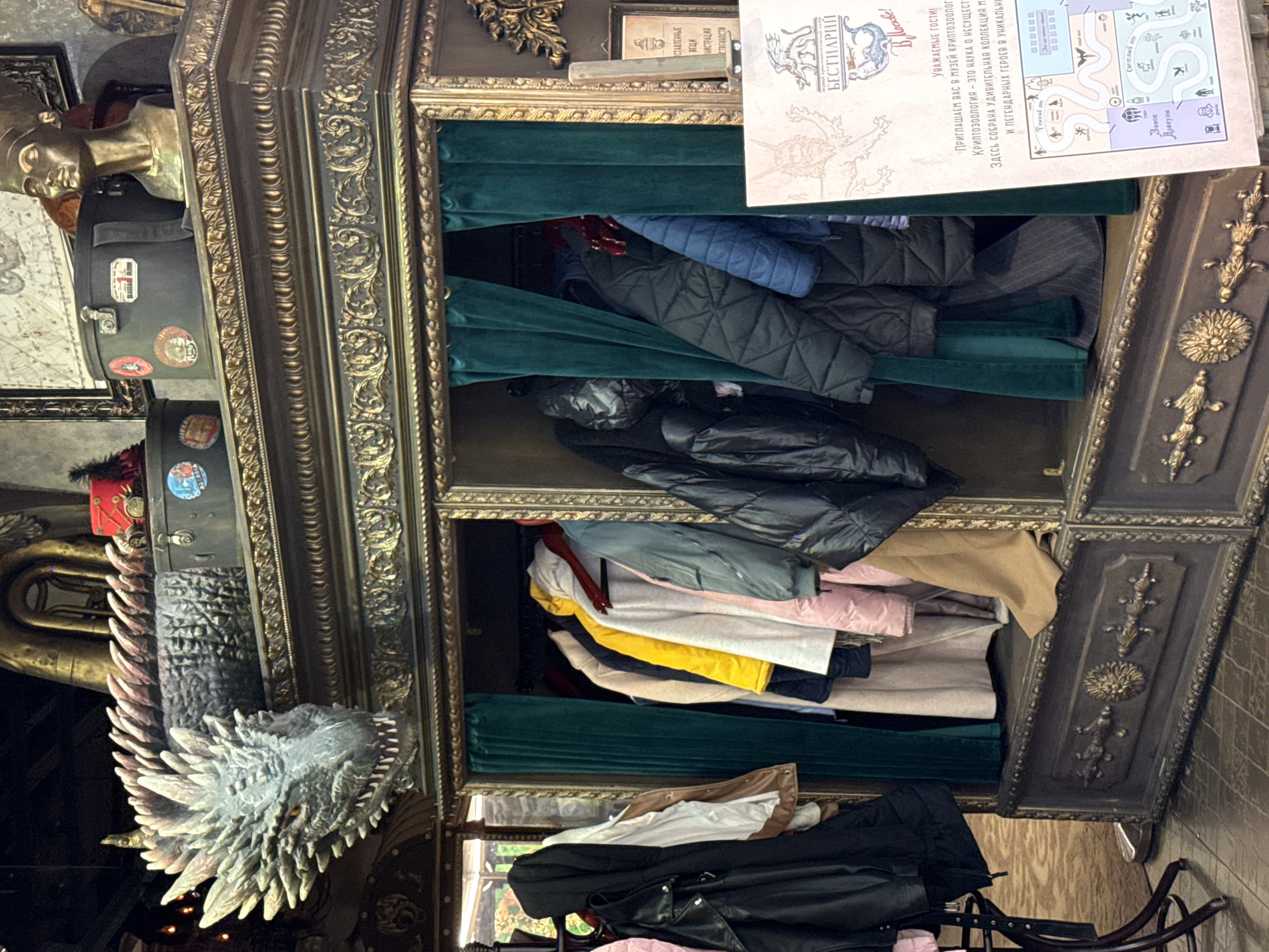 The image shows a richly decorated cabinet with multiple shelves, likely located in a museum. The cabinet is filled with folded clothing. A dragon head is mounted to the side of the cabinet, and a bust of what appears to be an Egyptian pharaoh is above it. A museum guide or map is partially obscuring the top shelf, indicating the cabinet might be within a museum of mythical or unusual creatures.
The text on the guide is in Russian. The text on the guide mentions “Museum of Cryptozoology”, suggesting it is located in Moscow, Russia.