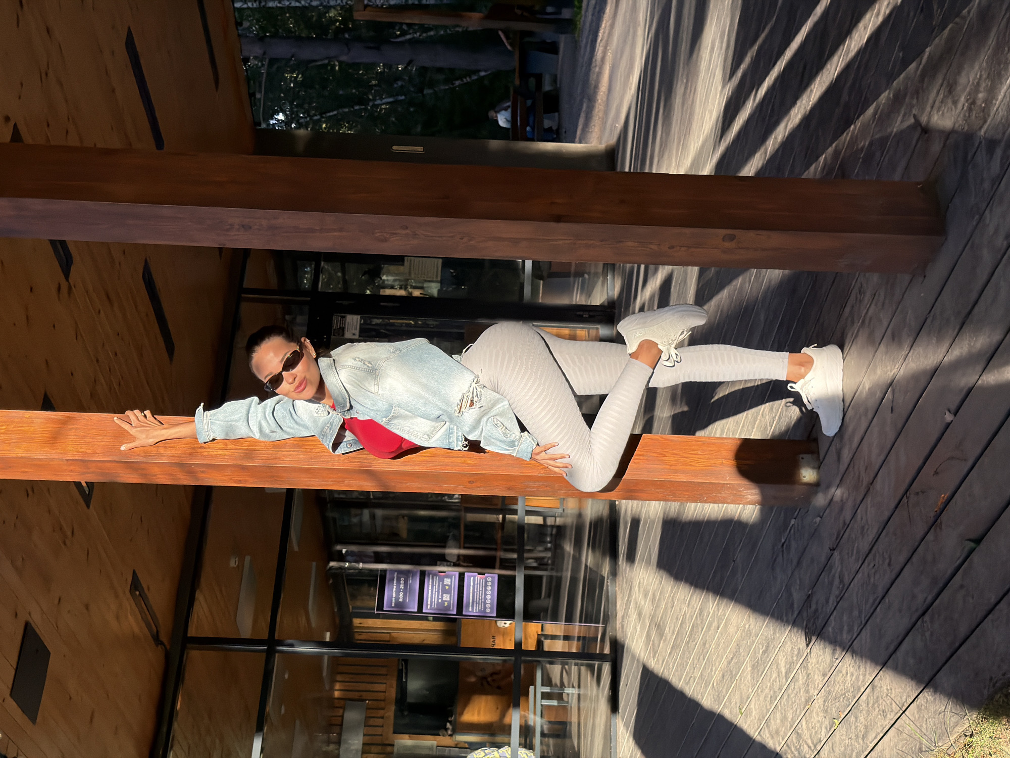 A young woman stands leaning against a wooden pillar outdoors. She is wearing a denim jacket, a red top, and light-colored leggings with a textured pattern. Her right arm is raised, touching the pillar, and her left leg is bent at the knee, with her foot resting on her thigh. She is looking to her left, wearing sunglasses. The background shows a building with large glass windows, a wooden deck, and trees. The scene is illuminated by sunlight, casting long shadows on the deck. The location is likely a park or outdoor recreational area within the city of Krasnoyarsk, Russia, given the architecture and surrounding trees.