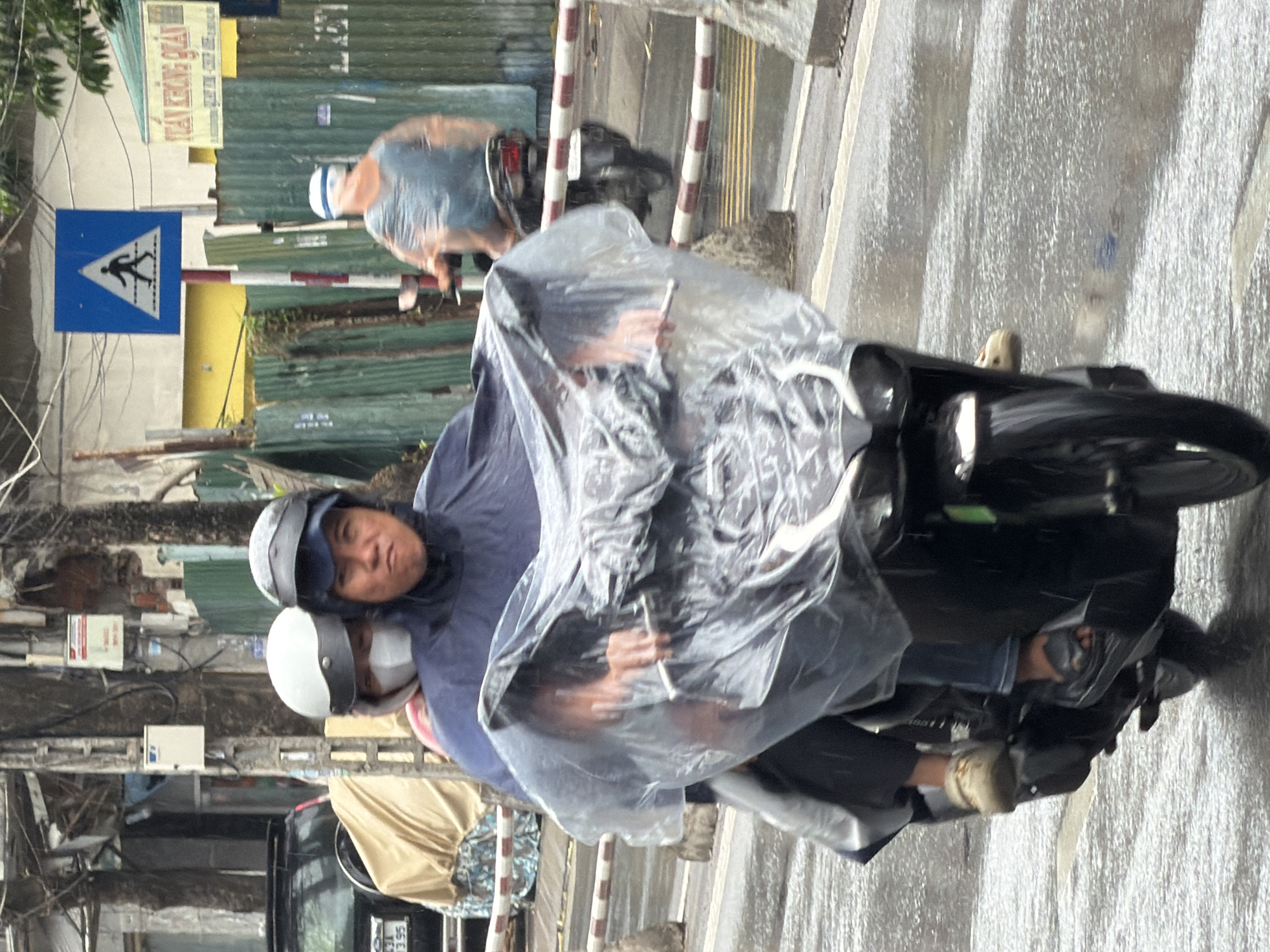 On a rainy day in Nha Trang, Vietnam, two individuals are riding a black scooter. The person in front, wearing a blue poncho and a black helmet, is driving. Behind them, a passenger wearing a white helmet and a white face mask is visible. Both are covered by a clear plastic poncho, making it difficult to see their clothing clearly. The road is wet, indicating recent rainfall. In the background, construction fencing and various signs are present, suggesting an urban environment undergoing development. A blue pedestrian crossing sign is prominent on the right side of the image. Another scooter with a rider in a transparent poncho is visible further down the road.