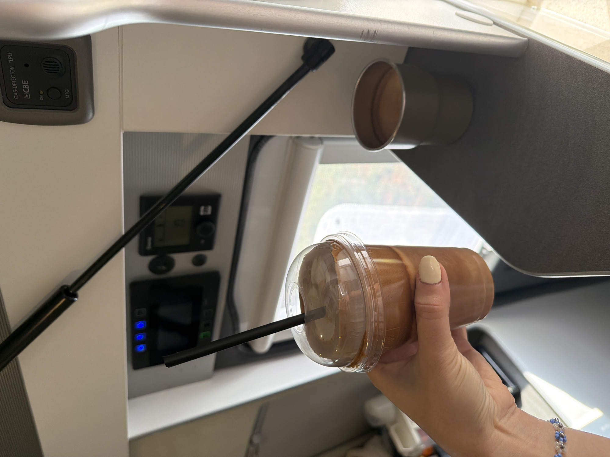 An interior view captures a hand holding a plastic cup filled with a brown beverage, complete with a black straw. Beside it, a small, olive-green cup sits on a dark surface. In the background, control panels with illuminated blue and green lights are visible. A gas detector is mounted on the wall above these panels. The scene appears to be within a vehicle or stationary dwelling, possibly a caravan or train compartment, given the presence of control interfaces and a window in the background. The overall ambiance is functional and personal, with the beverage suggesting a moment of relaxation or transit. There is no indication of the city of Sarnitsa, Bulgaria.