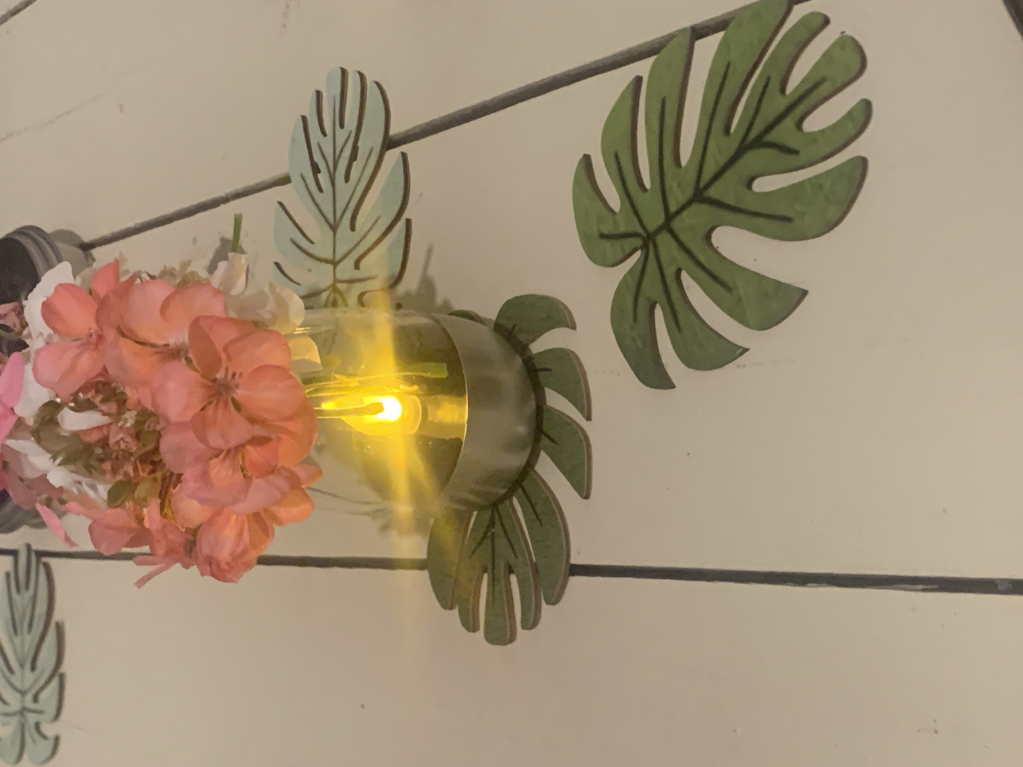 This media file displays a close-up of a decorative light fixture mounted on a light-colored, horizontally paneled wall. The fixture features a glass bulb emitting a warm yellow light, wrapped in a metal band. Adorning the fixture are decorative elements resembling monstera leaves, painted in shades of green and light blue. A cluster of pink and white flowers is positioned to the left of the light bulb, some appearing to be hydrangeas. The background consists of the textured wall, with a dark horizontal line running across it, possibly a shelf or part of the fixture's mounting. The lighting suggests an indoor setting, potentially a living space or a decorative display area. The overall aesthetic is rustic and bohemian, with a focus on naturalistic decorative elements. No people are visible, and no text is present in the image. The image is cropped to focus on the light fixture and its immediate surroundings, obscuring any wider view of the location.