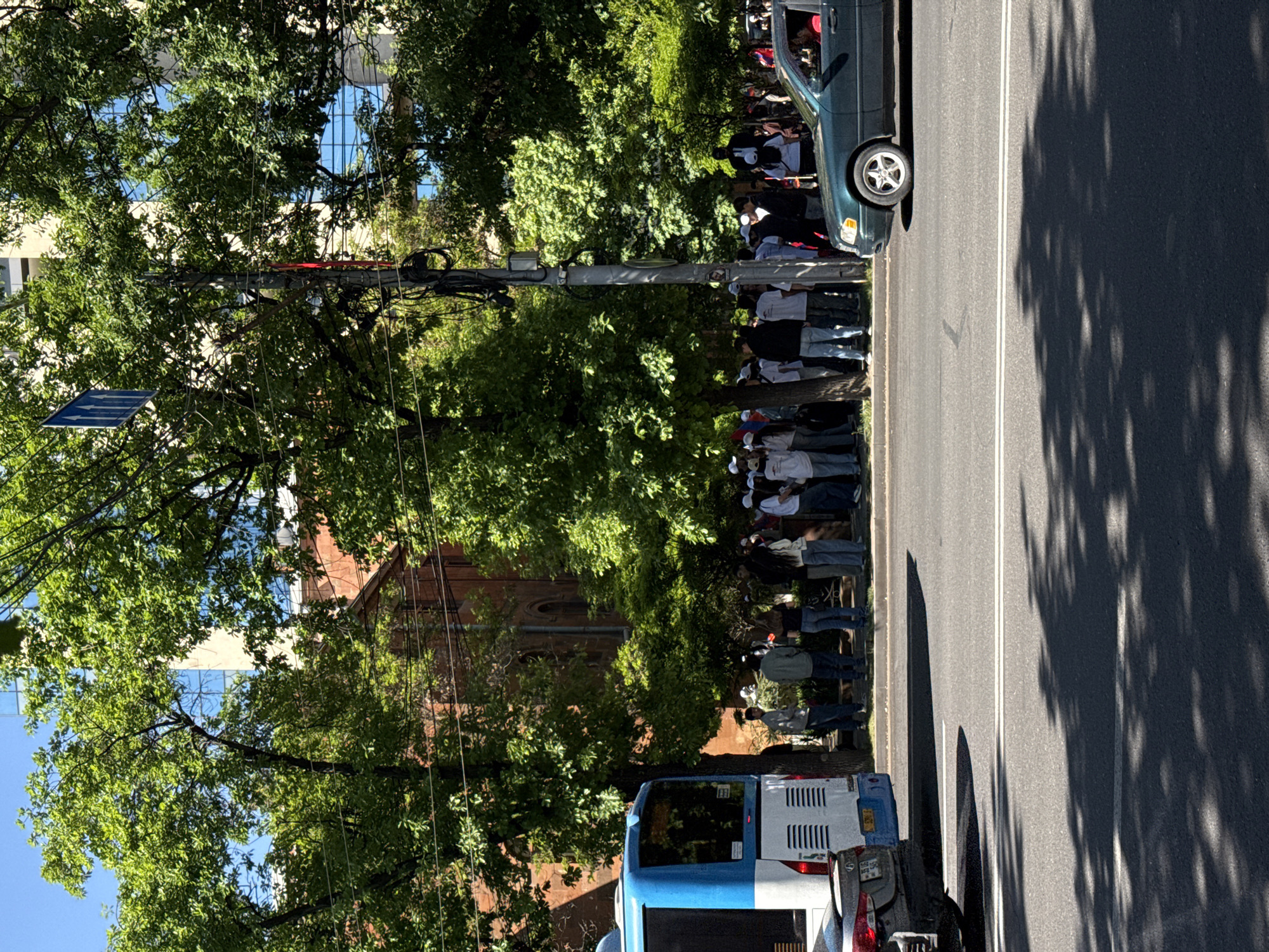 The image shows a street in Yerevan, Armenia, with people lined up along the sidewalk next to a road with cars and buses. Trees line the sidewalk and cast shadows on the road. Buildings are visible in the background, including one with a red brick facade and another with a blue glass facade. A traffic sign is attached to a pole with many hanging wires, which are strung throughout the tree branches.