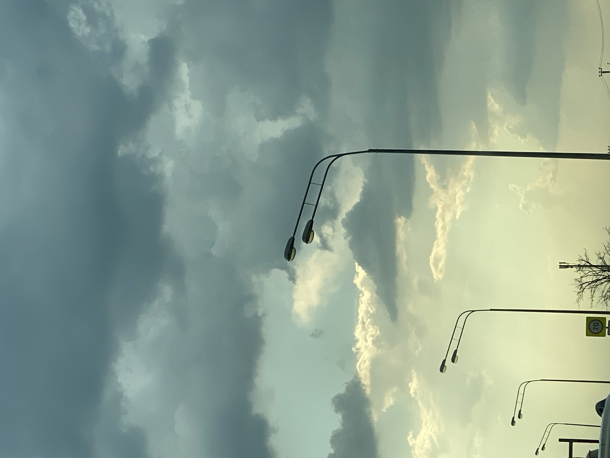The image depicts a moody sky with dark, heavy clouds dominating the upper half, transitioning to lighter, brighter clouds and a pale yellow-green sky towards the bottom. Silhouetted against this sky are several streetlights. The most prominent streetlight, located on the right side, is tall with a single pole and two lamp fixtures at the top, angled outwards. To the left and further back, there are two more streetlights, each with a single pole and two lamp fixtures. In the lower portion of the image, a portion of a white vehicle and a yellow speed limit sign with the number "70" are visible. A bare-branched tree and a communication tower can also be seen in the distance. The scene appears to be on the outskirts of a city, possibly along a road or highway, given the presence of streetlights and the speed limit sign.