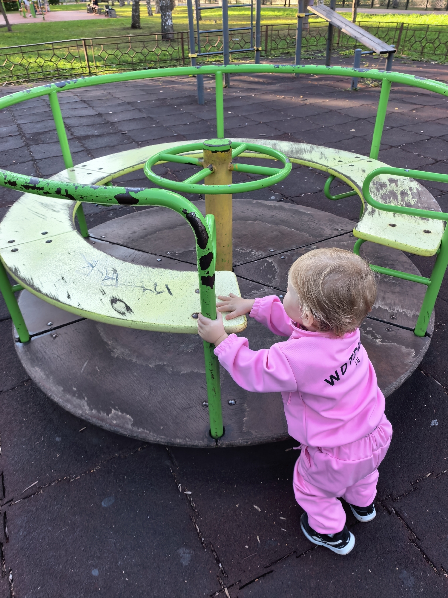 The image depicts a young child, seen from behind, standing on a playground merry-go-round. The child is wearing a pink tracksuit and black and white sneakers. Their hands are resting on the edge of the merry-go-round, which is painted bright green and yellow and shows signs of wear. The merry-go-round itself is situated on a dark, rubberized playground surface, characteristic of modern play areas. In the background, a grassy area with trees and a metal fence is visible, suggesting the location is a park or public recreational space. The lighting indicates it is daytime, possibly late afternoon given the warm tone of the light on the grass. No specific landmarks or signs are visible that would definitively place the location within Sterlitamak, Russia, beyond the provided prompt.