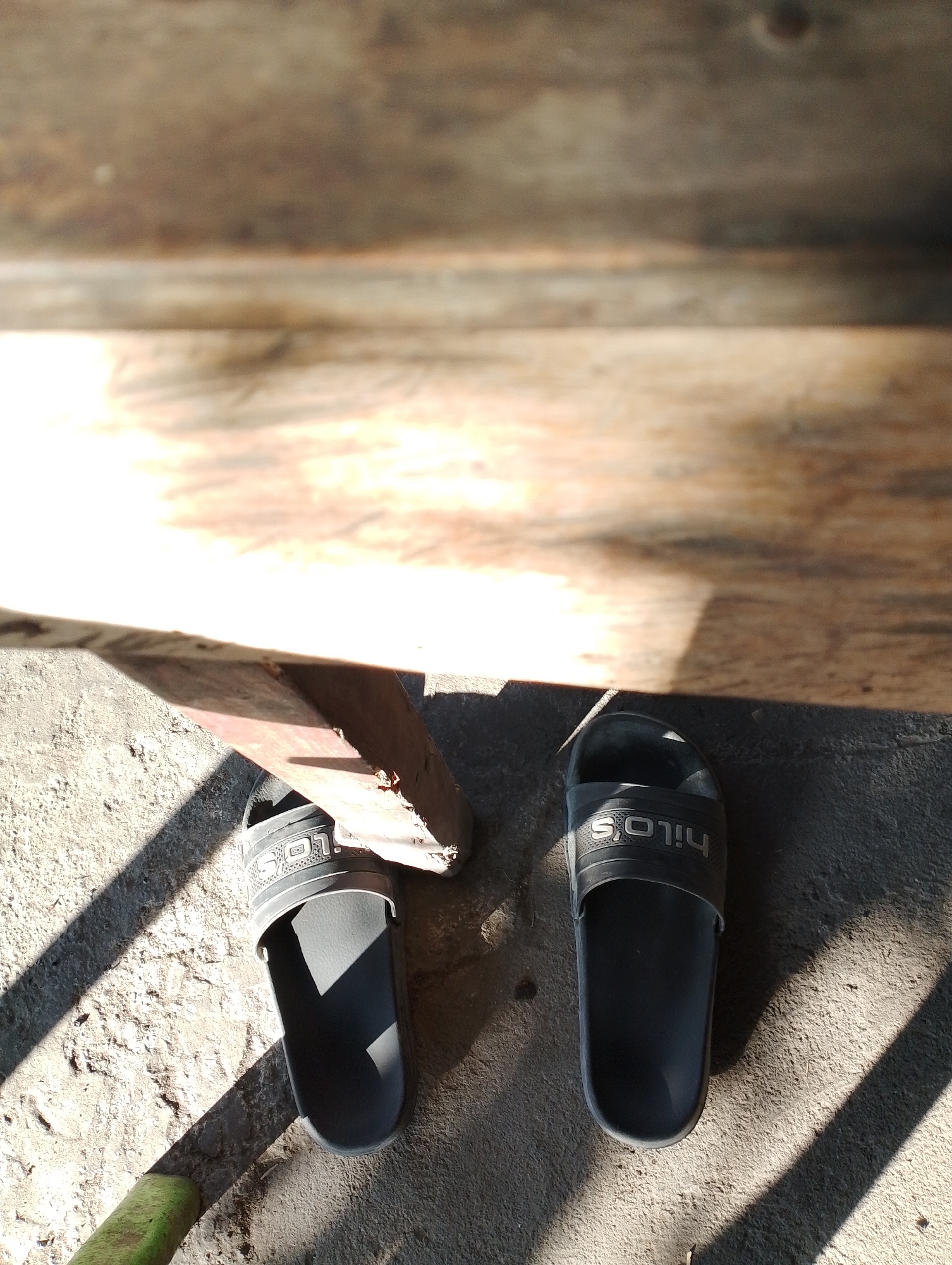 Two black Hilo's brand slides are positioned on a textured concrete surface. The slides are arranged side-by-side, with the left slide slightly angled towards the viewer and the right slide facing directly forward. The upper strap of both slides features the embossed Hilo's logo. The concrete is illuminated by dappled sunlight, creating distinct striped shadows across the ground. In the background, a wooden surface is visible, appearing weathered and aged. A portion of a metal support structure, possibly a part of a wooden platform or bench, is also present between the slides, casting a shadow. A hint of a green object, possibly a tool or piece of debris, is visible at the bottom left corner of the image. The overall scene suggests an outdoor setting, likely a residential area or a simple outdoor structure, such as a porch or walkway, in Jambean, Indonesia.