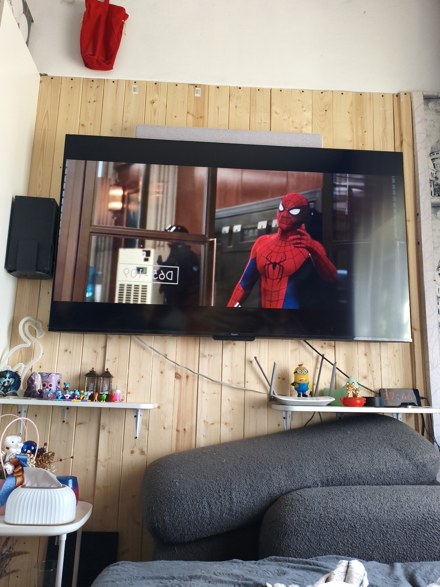 This image depicts an indoor scene, likely a living room, with a large television screen displaying a movie featuring Spider-Man. The television is mounted on a wall paneled with light-colored wood. To the left of the television, a black speaker is attached to the wall. Below the television, on a white shelf, are various decorative items, including small figurines, lanterns, and what appears to be a tissue box. A plush gray sofa is partially visible in the foreground. Above the television, a red fabric bag is hanging from the wall. The overall impression is of a cozy, lived-in space. There is no information to indicate the location is in Vladivostok, Russia.