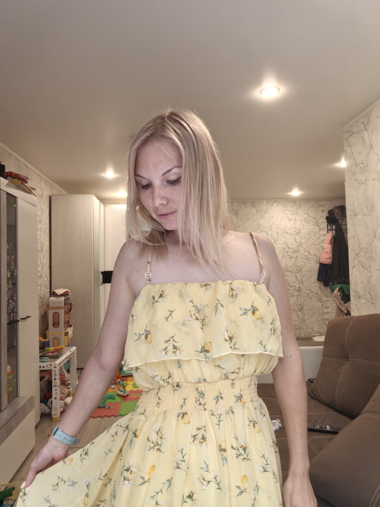The image depicts an interior scene, likely a living room or bedroom, with a woman in the foreground. She has blonde hair and is wearing a yellow sundress with a floral and lemon print. Her gaze is directed downwards. The background reveals elements of a domestic environment, including furniture such as a sofa and cabinets, as well as children's toys on a play mat. The lighting suggests indoor illumination. There is no visible information or indication that this scene is located in the city of Sterlitamak, Russia.