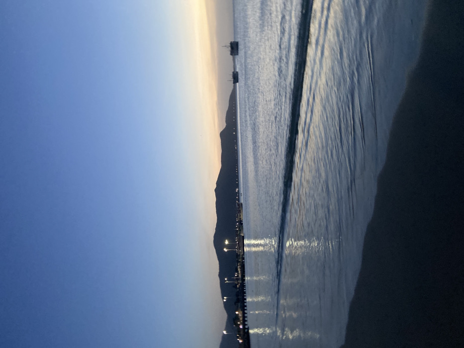 A landscape shot of a beach at dusk. Mountains rise in the distance, their dark silhouettes contrasting with the fading sunlight on the horizon. Bright lights line the shore. Gentle waves lap the sandy beach. The sky transitions from a deep blue to a soft yellow-orange, mirroring the light reflecting on the ocean's surface. Two structures are offshore, also silhouetted against the light. The sand closer to the viewer is darker and indistinct.