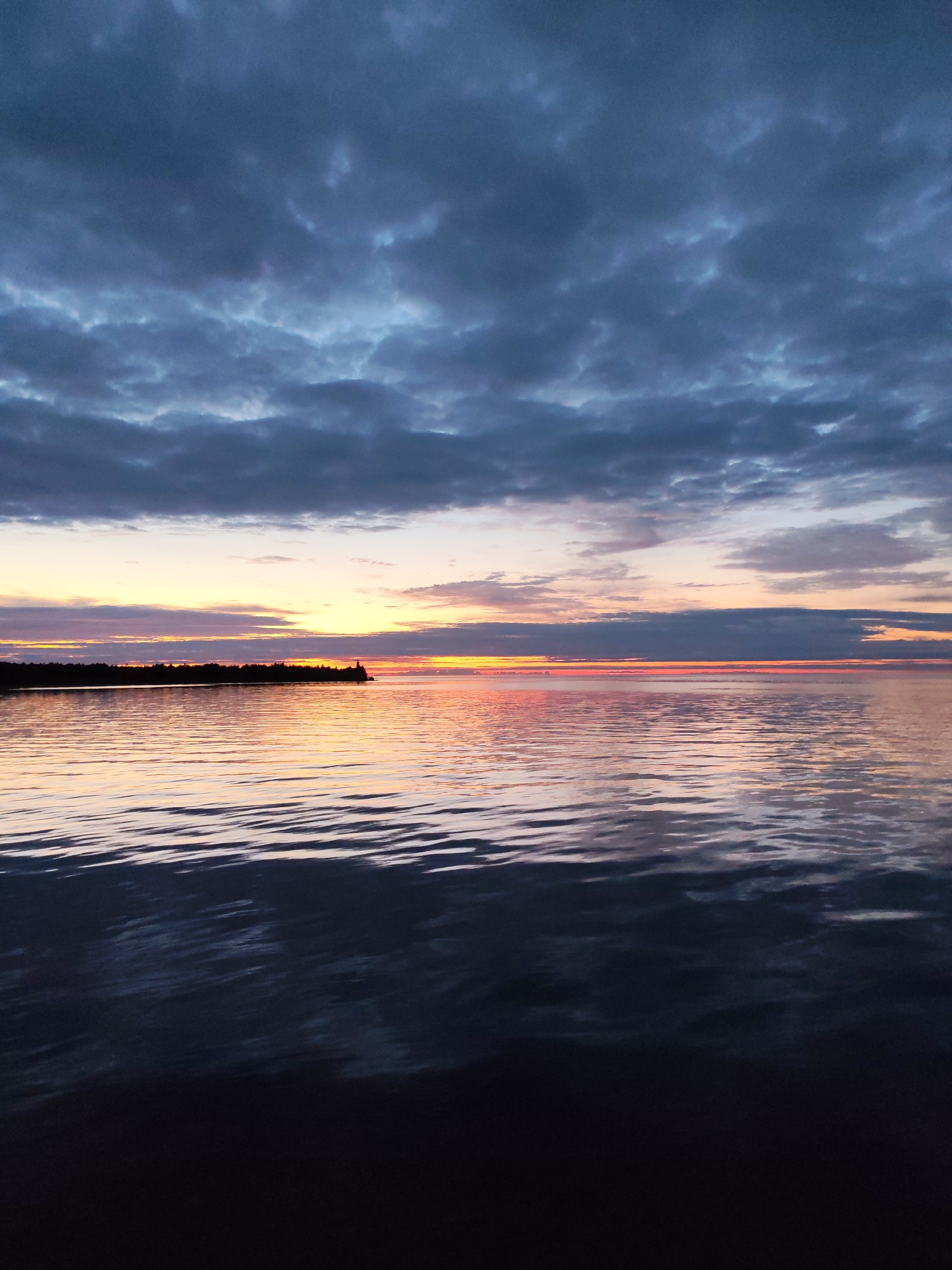 The image depicts a serene natural landscape at dusk. The sky is filled with dramatic, dark grey clouds, interspersed with lighter patches that suggest residual sunlight. Below the clouds, a band of soft, warm colors – pinks, oranges, and yellows – stretches across the horizon, indicating a sunset. This colorful glow is reflected in the calm surface of a body of water, which occupies the lower half of the frame. The water itself is dark, with subtle ripples creating patterns of light and shadow. In the distance, a dark silhouette of a treeline stretches along the horizon, with a small, lighter structure, possibly a lighthouse or a tower, visible on a small peninsula jutting out into the water. The overall impression is one of tranquility and the beauty of nature during the twilight hours. The location of Shcheleiki, Russia is not discernible from the visual information provided in the image.