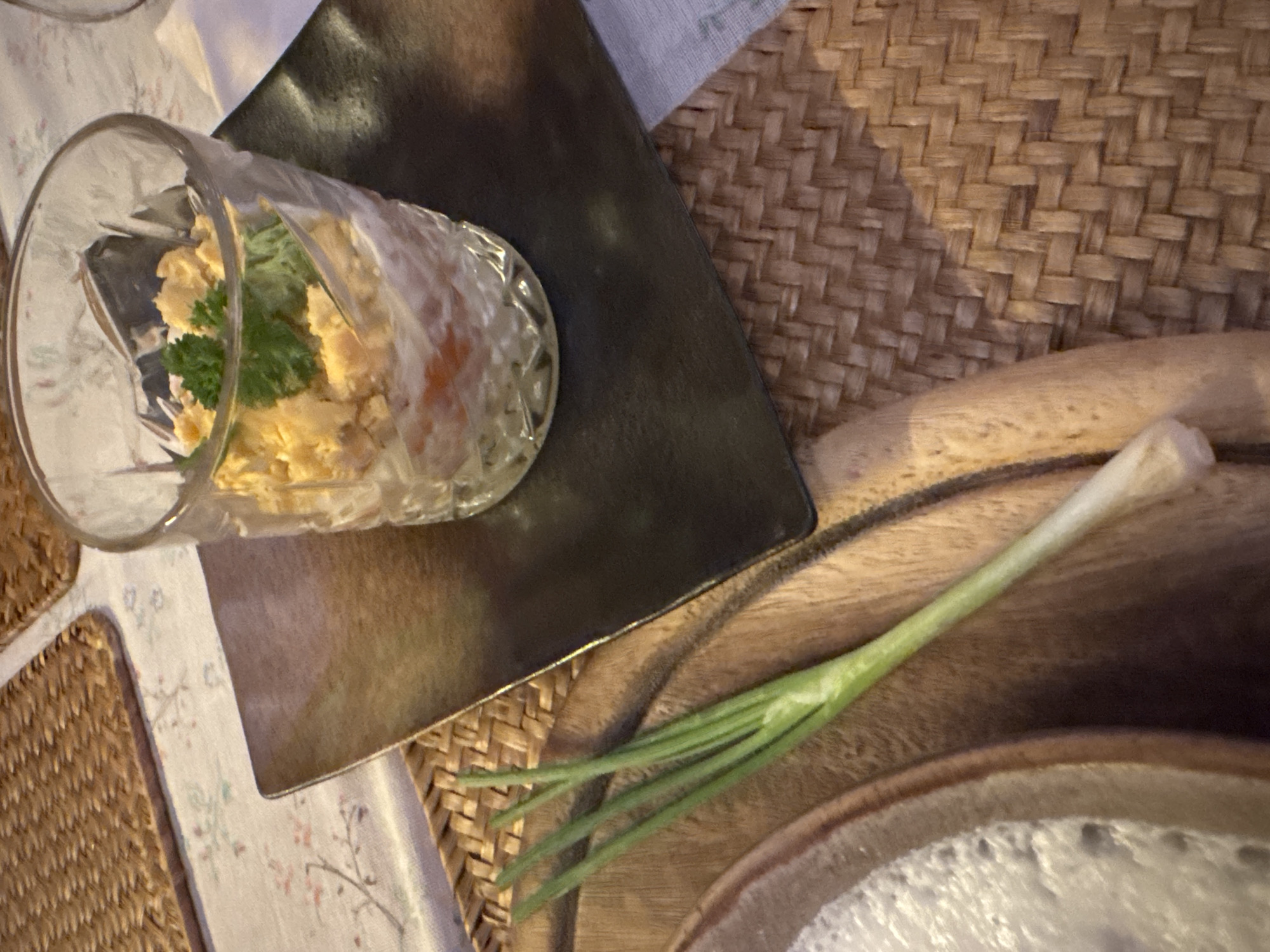 This is a close-up, top-down shot of a table setting. The main subjects are a clear glass containing a food item, a dark square plate, and a green onion. The food in the glass appears to be a creamy mixture, possibly a salad or dip, with a sprig of parsley or lettuce on top. The glass is placed on the dark plate, which in turn is situated on a woven placemat. The green onion lies diagonally across a wooden serving platter in the foreground. The lighting suggests it could be evening or an indoor setting with artificial light, as there are noticeable shadows. The overall mood is calm and domestic, possibly indicating a meal in preparation or just served. There is no visible text in the image.