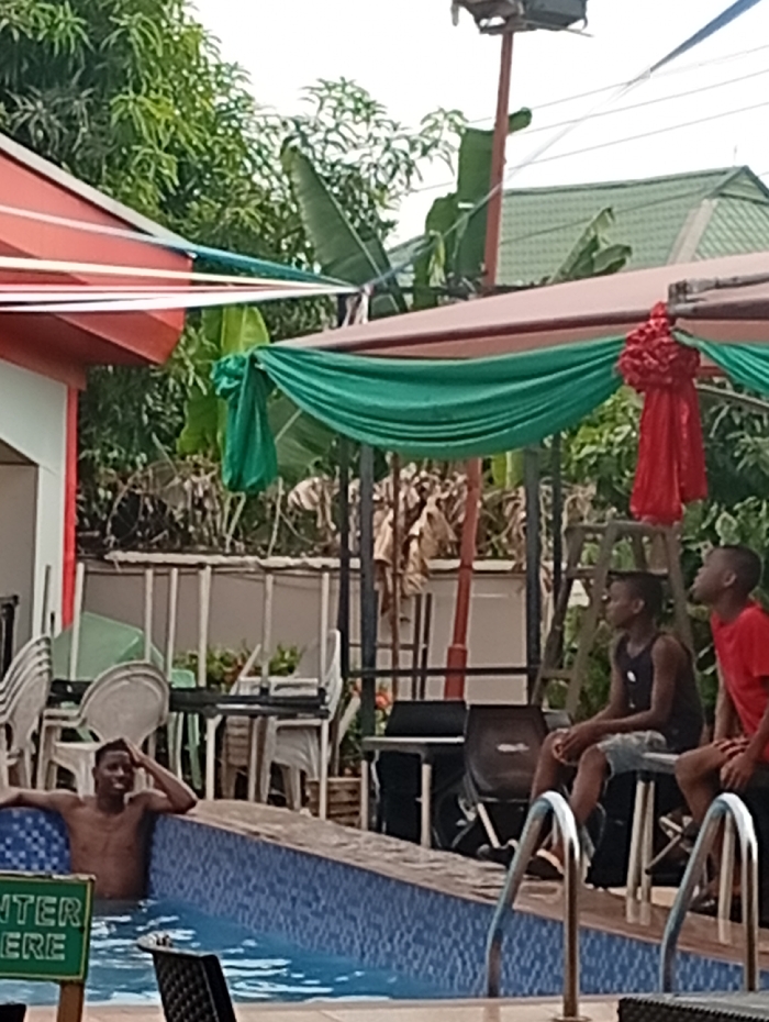 The image captures an outdoor leisure scene at what appears to be a poolside area within the Akpaka Forest Reserve, Nigeria. In the foreground, a swimming pool with blue mosaic tiles is visible, featuring metal steps on the right.
One young man is partially submerged in the pool, looking towards the right with a smile on his face and his hands near his head, indicating relaxation or enjoyment. To his right, two other young males are seated on the edge of the pool deck or on stools. The one closer to the viewer wears a dark tank top and shorts, while the other is in a red t-shirt; both are looking towards the right side of the frame.
The background shows a lively setting with several structures and lush vegetation. A building with a red roof and white and red striped decorations is on the left. A shaded area stretches across the mid-ground, adorned with draped green fabric and a prominent red, possibly floral, decoration, suggesting a festive or event-oriented atmosphere. Numerous white plastic chairs are arranged or stacked in the background, further hinting at a gathering. A building with a green roof is visible in the distance, partially obscured by trees and other structures. Green trees and foliage are abundant throughout the scene, contributing to the outdoor ambiance.
The time of day appears to be bright daylight, suggesting a clear or partly cloudy sky. The overall mood conveyed is one of relaxation and social activity.
A partially visible green sign near the pool reads "INTER HERE" or "ENTER HERE".