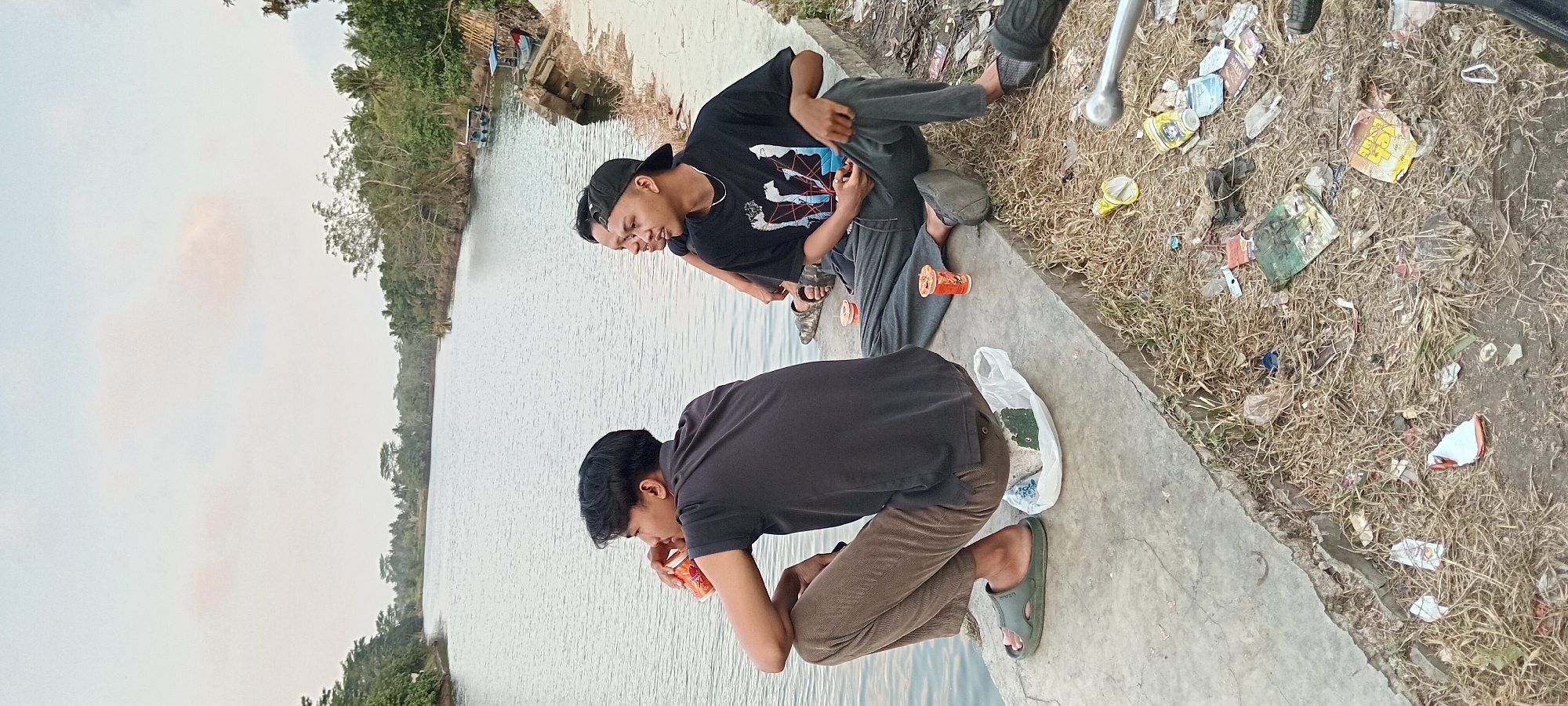 Three young men are sitting on a concrete embankment next to a body of water, likely a river or canal, in Kalipucang, Indonesia. The man on the left is wearing a dark gray polo shirt and brown pants, and is holding a cup to his mouth. He is wearing green sandals. In front of him is a white plastic bag. The man in the middle is wearing a black t-shirt with a graphic print and dark gray sweatpants. He is wearing black slip-on shoes. The man on the right is wearing a black t-shirt and dark gray sweatpants, and has a black baseball cap on backwards. He is also wearing black slip-on shoes. All three men are looking towards the water. Two orange cups are visible on the embankment between the men. The ground next to the embankment is covered in dry grass and debris, including various pieces of trash. In the background, the water stretches into the distance, lined with trees and vegetation. The sky is overcast.