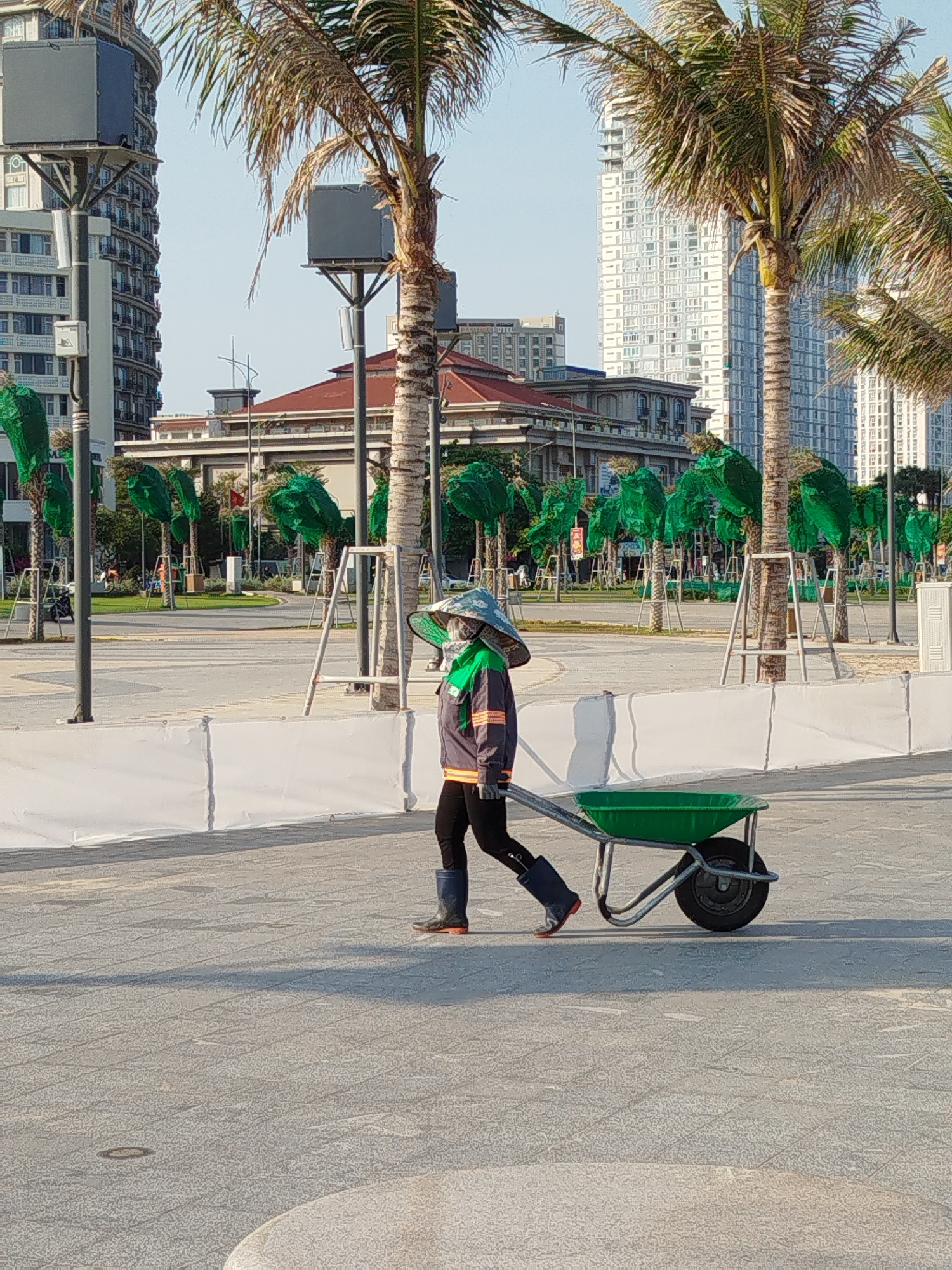 A worker wearing protective clothing is walking and pulling a green wheelbarrow across a paved area in Ho Chi Minh City, Vietnam. The worker is wearing a wide-brimmed hat, face covering, dark clothing, a gray and orange striped jacket, and boots. Rows of trees covered in green mesh line the background, and buildings are visible in the distance. A temporary white barrier is present. The day appears sunny.