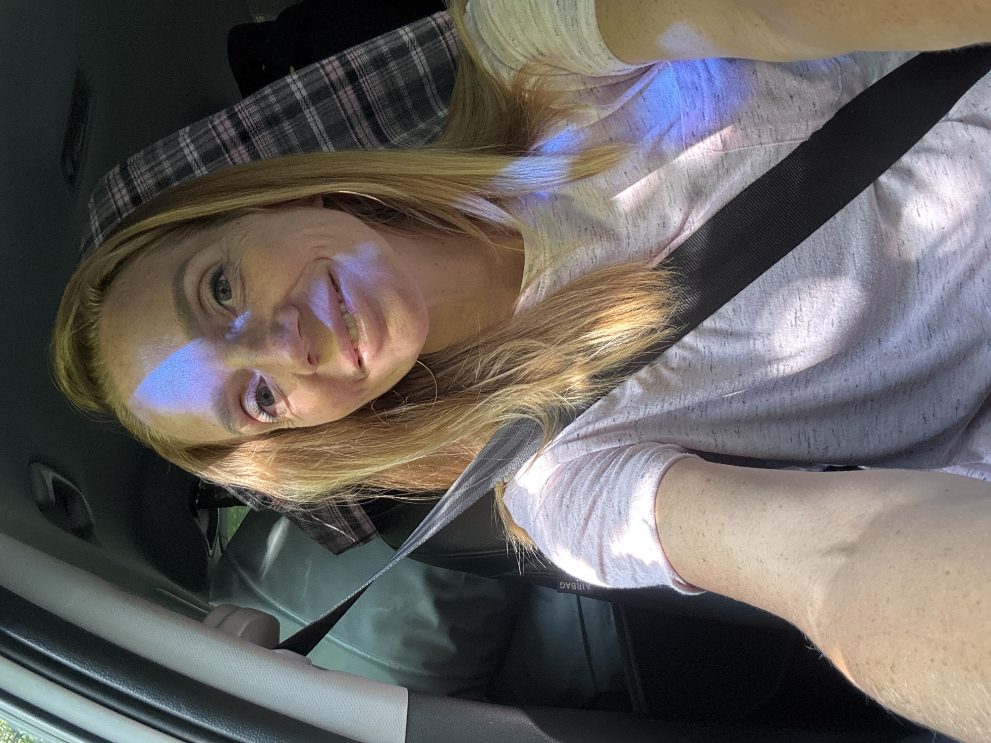 A woman is sitting in the passenger seat of a car, wearing a pink shirt and a seatbelt. There is a plaid blanket behind her. She has blonde hair and is smiling at the camera. The car appears to be stationary. The image was taken in Portovoe, Russia.