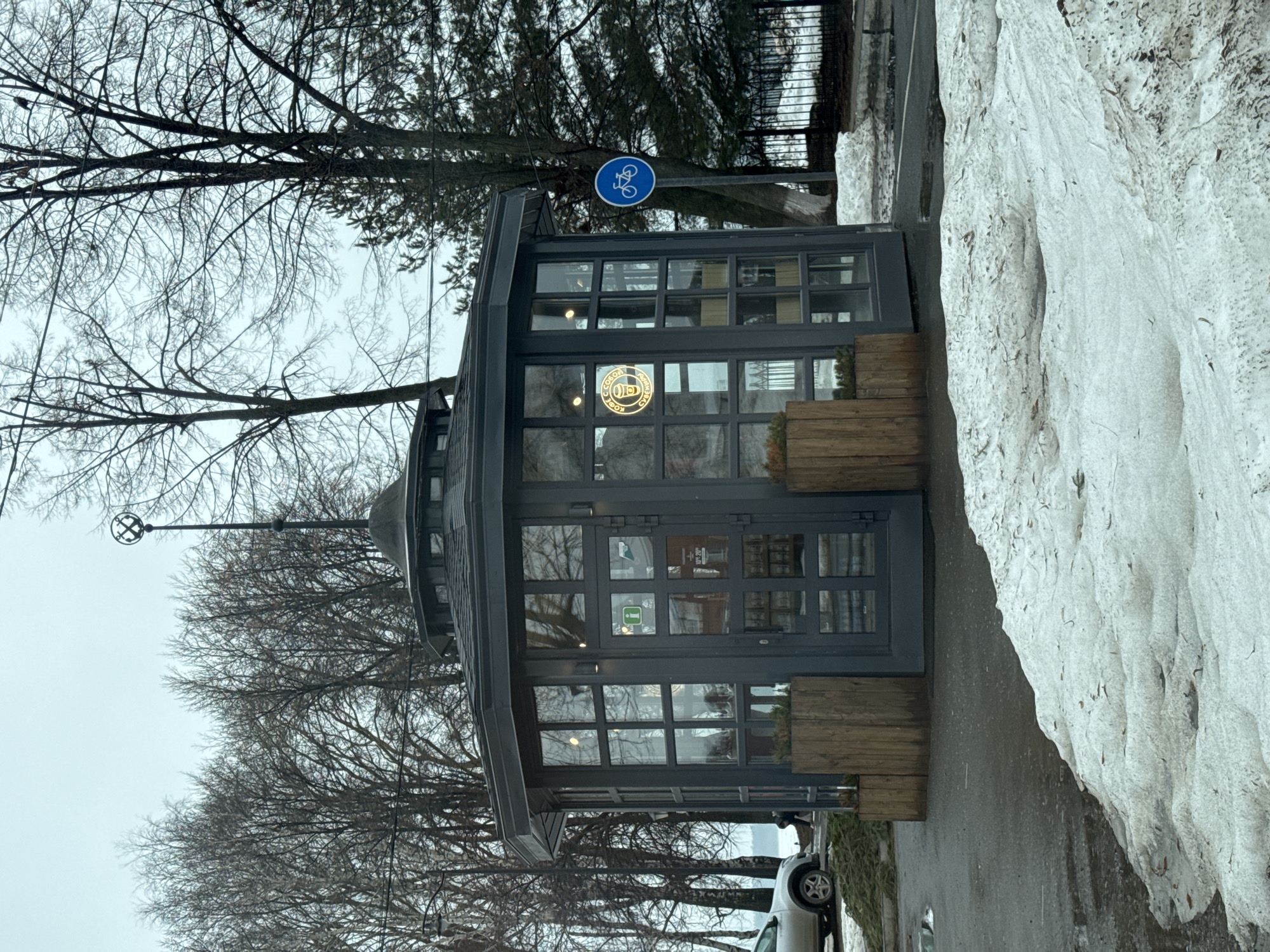 The image captures a small cafe in Votkinsk, Russia. The cafe is a dark-colored structure with a hexagonal shape, enclosed by large windows framed with dark wood. The windows display a circular, illuminated logo. Potted greenery lines the bottom of the cafe's windows. A round, blue bicycle sign is posted. A layer of melting snow borders the street next to the cafe. Bare trees surround the cafe. A streetlamp and parked cars are also visible.