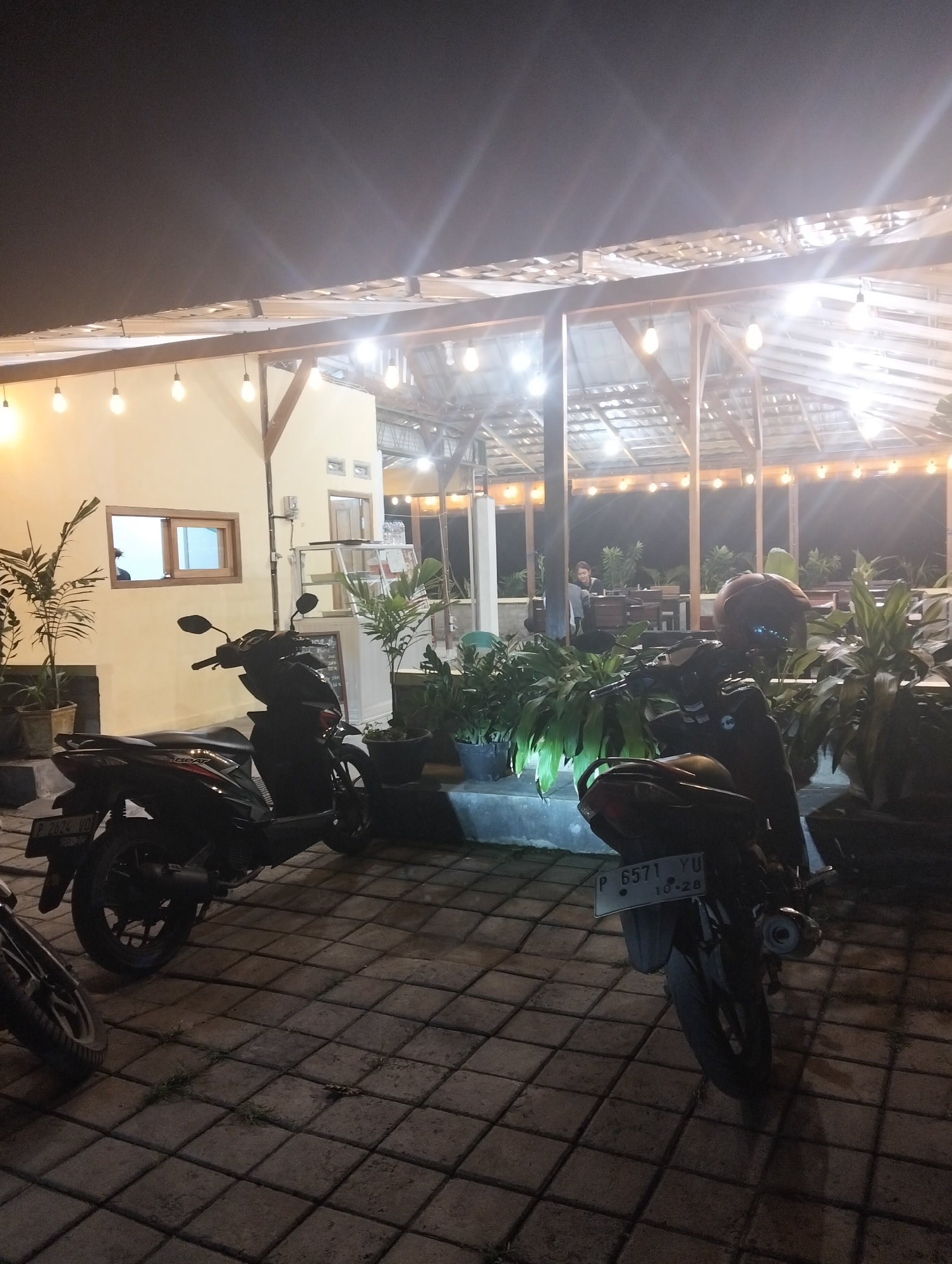 The image depicts an outdoor seating area at night, likely a cafe or restaurant, with a thatched roof supported by wooden beams and illuminated by strings of lights. Paved square tiles form the ground. Two motorcycles are parked in the foreground; one is black with a license plate reading "P 2624 VD", and the other is partially visible to the right. A person is seated at a table inside the establishment, and a woman is visible through a window in the building on the left. The overall ambiance is dimly lit and relaxed.