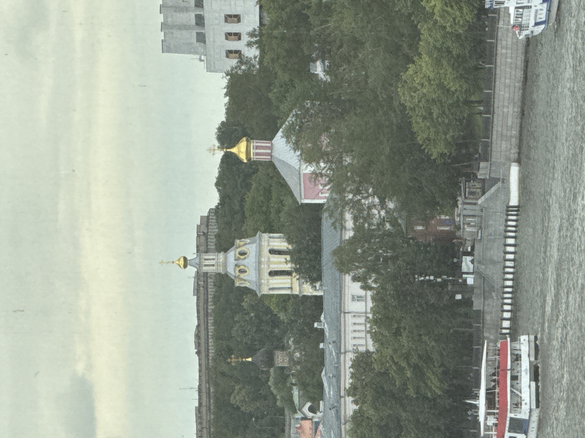This is a vertically oriented, slightly blurry, wide-angle photograph of a cityscape bordering a body of water. The scene appears to be illuminated by soft, diffused light, suggesting an overcast day or perhaps early morning or late afternoon.
The primary subjects are religious structures with gold domes, characteristic of Russian Orthodox churches. One prominent church, with a yellow bell tower topped with a golden dome and cross, is partially obscured by dense green trees. Another smaller, dark dome with a golden cross is visible further back. Adjacent to the religious buildings are modern, multi-story structures, one of which is light gray with a simple, blocky design and visible windows.
Along the right side of the image is a wide, grey body of water, likely a river, with a textured, rippling surface. A paved embankment runs along the water's edge, with steps leading down to the water in places. Several boats are visible docked or moving in the water. One is a white passenger boat with red accents, carrying several visible people on its deck. Another white boat, possibly a ferry, is docked further up the embankment.
The overall setting suggests a historical and modern urban environment coexisting, with a focus on the architecture of religious buildings and the presence of a major waterway. The dense foliage indicates a time of year with active tree growth, likely spring or summer. The mood is calm and somewhat muted, likely due to the overcast weather. No visible text is present in the image.